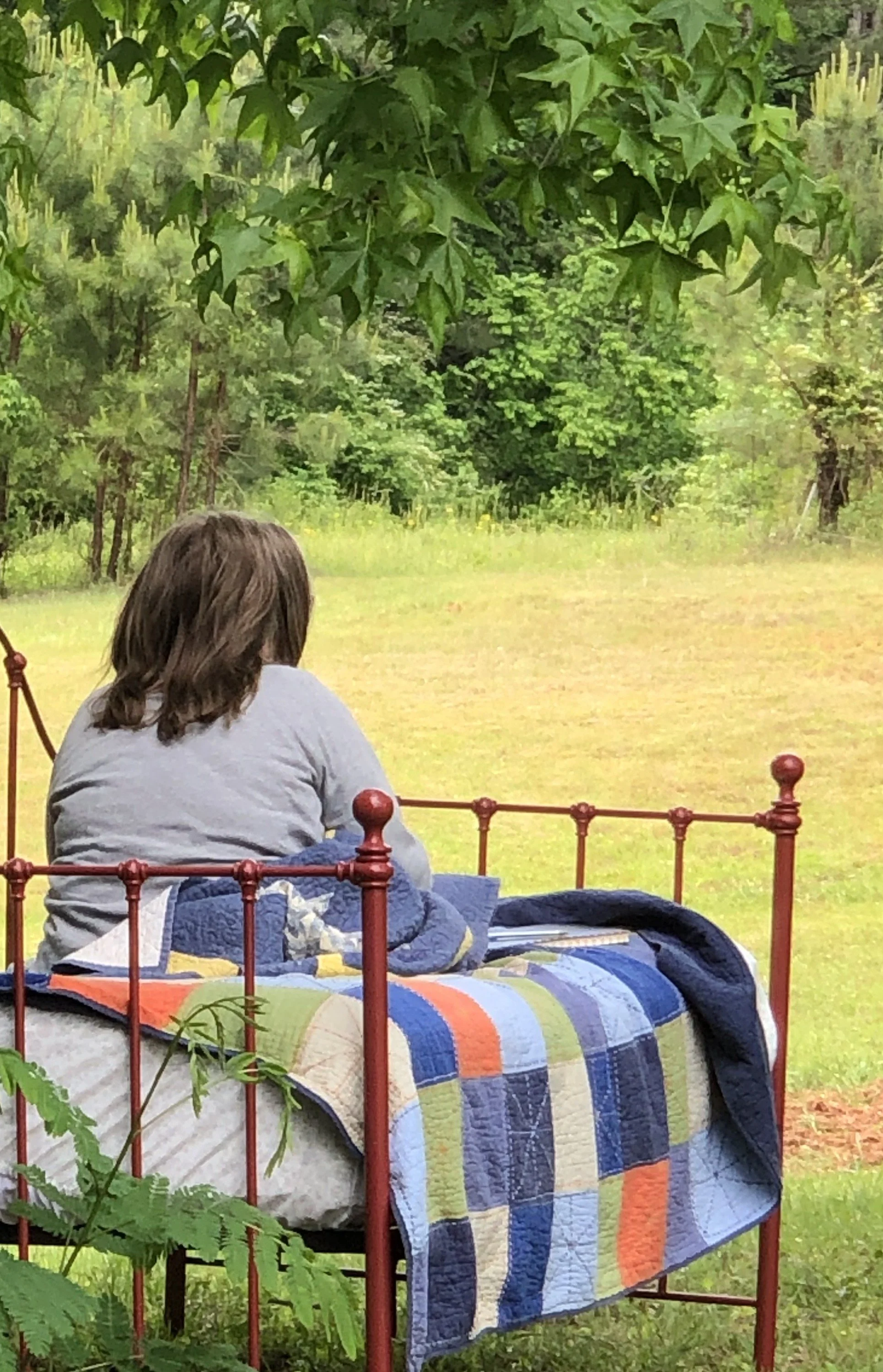 A person with brown hair sitting on a bed with a colorful striped quilt, facing away, outdoors in a lush green garden or park. A wooden fence and trees are visible in the background.