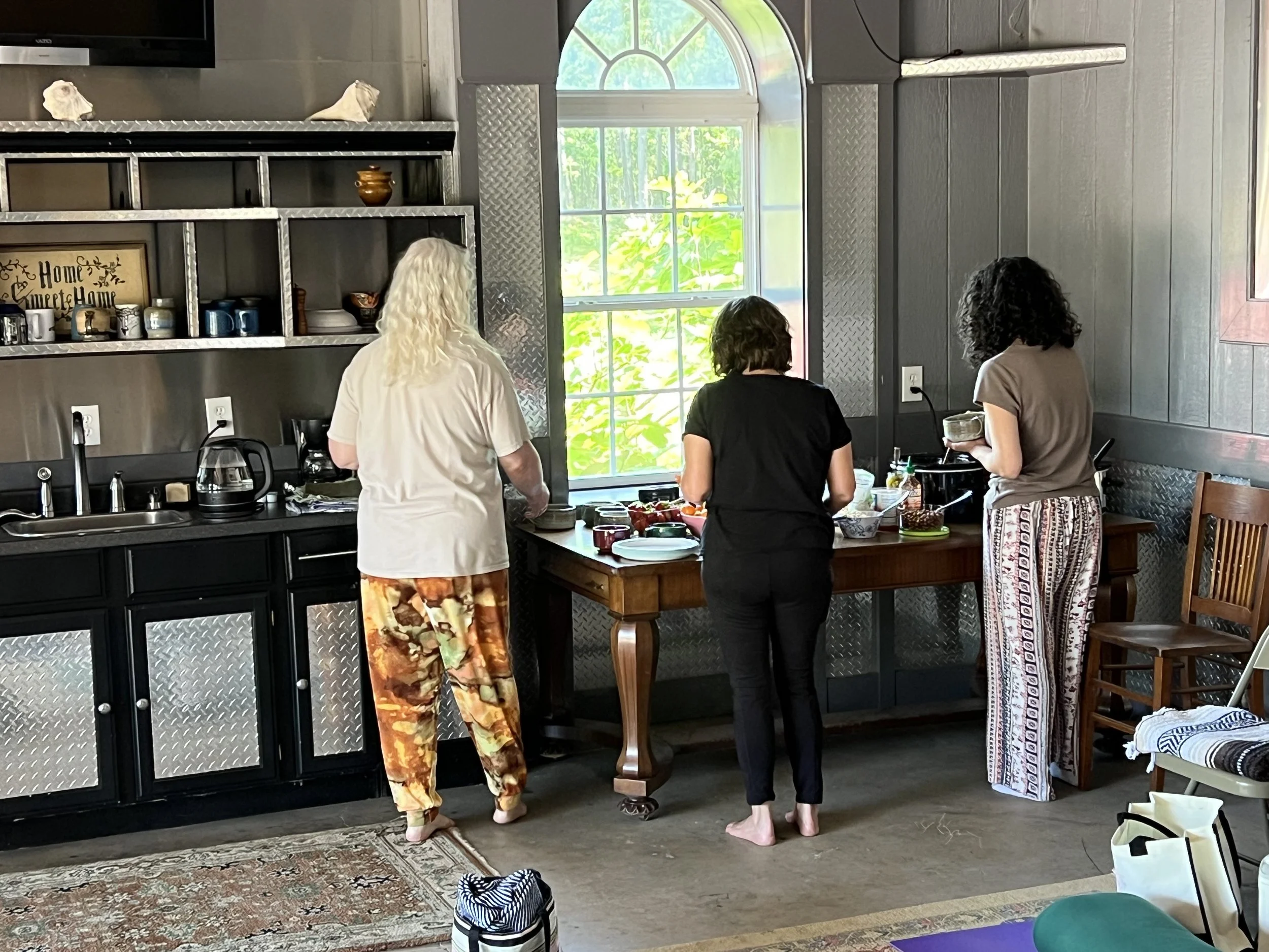 Three women are preparing food at a kitchen counter near a large window with lush green foliage outside. The women are standing barefoot, and various kitchen items such as plates, bowls, and utensils are visible on the counter.
