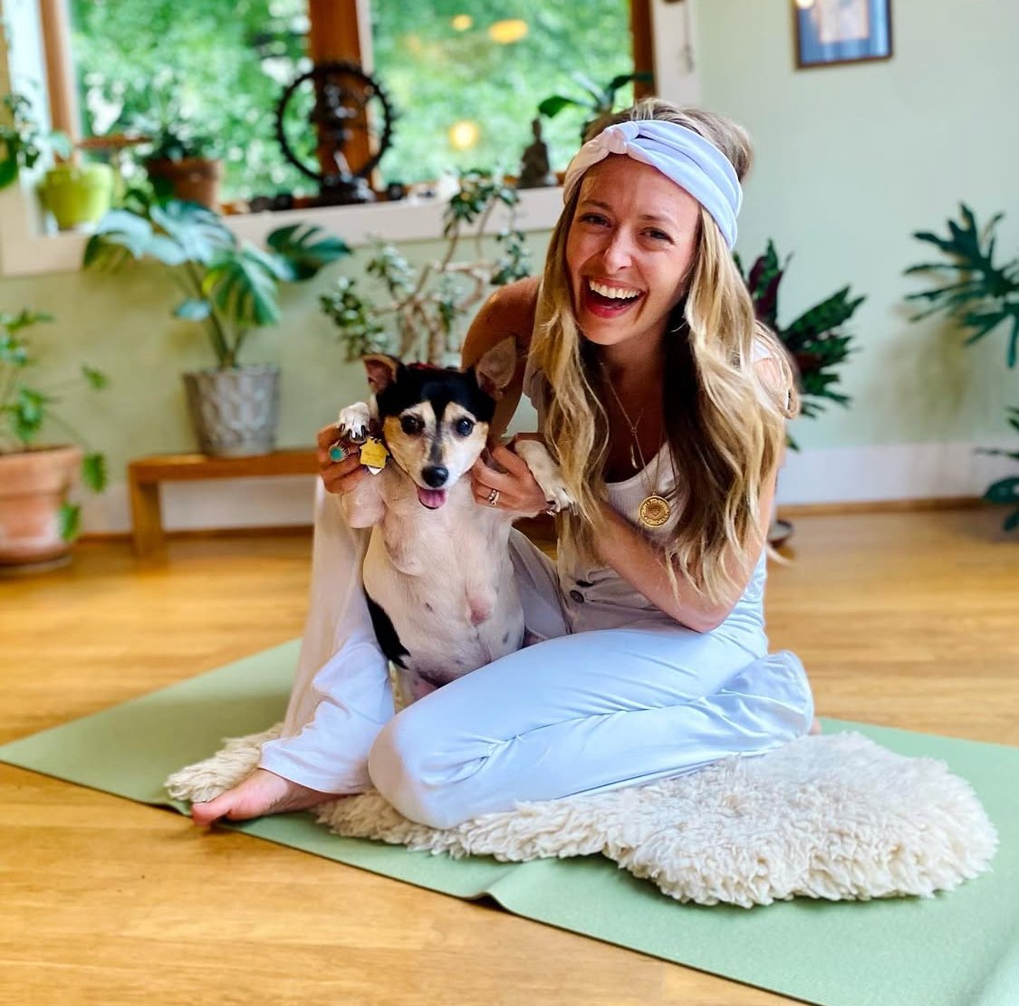A woman with long blonde hair, wearing a light blue headband and white outfit, smiling and kneeling on a yoga mat. She is holding a small black and tan dog with pointy ears and a collar. There are green plants and wooden furniture in the background.