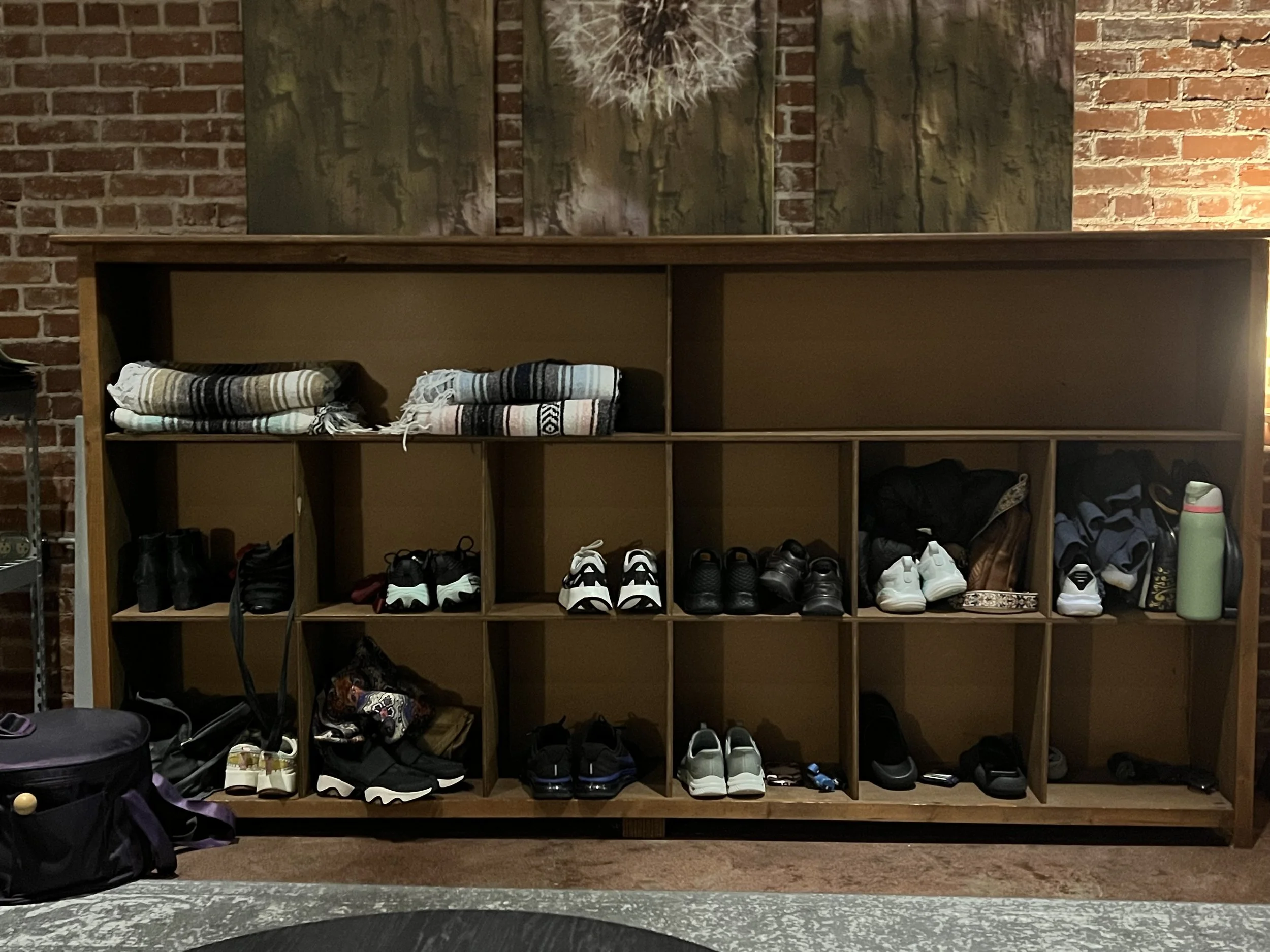 A wooden cubby shelf against a brick wall, filled with shoes, bags, and folded blankets on top, with additional items on the floor including a backpack and a purple bag.