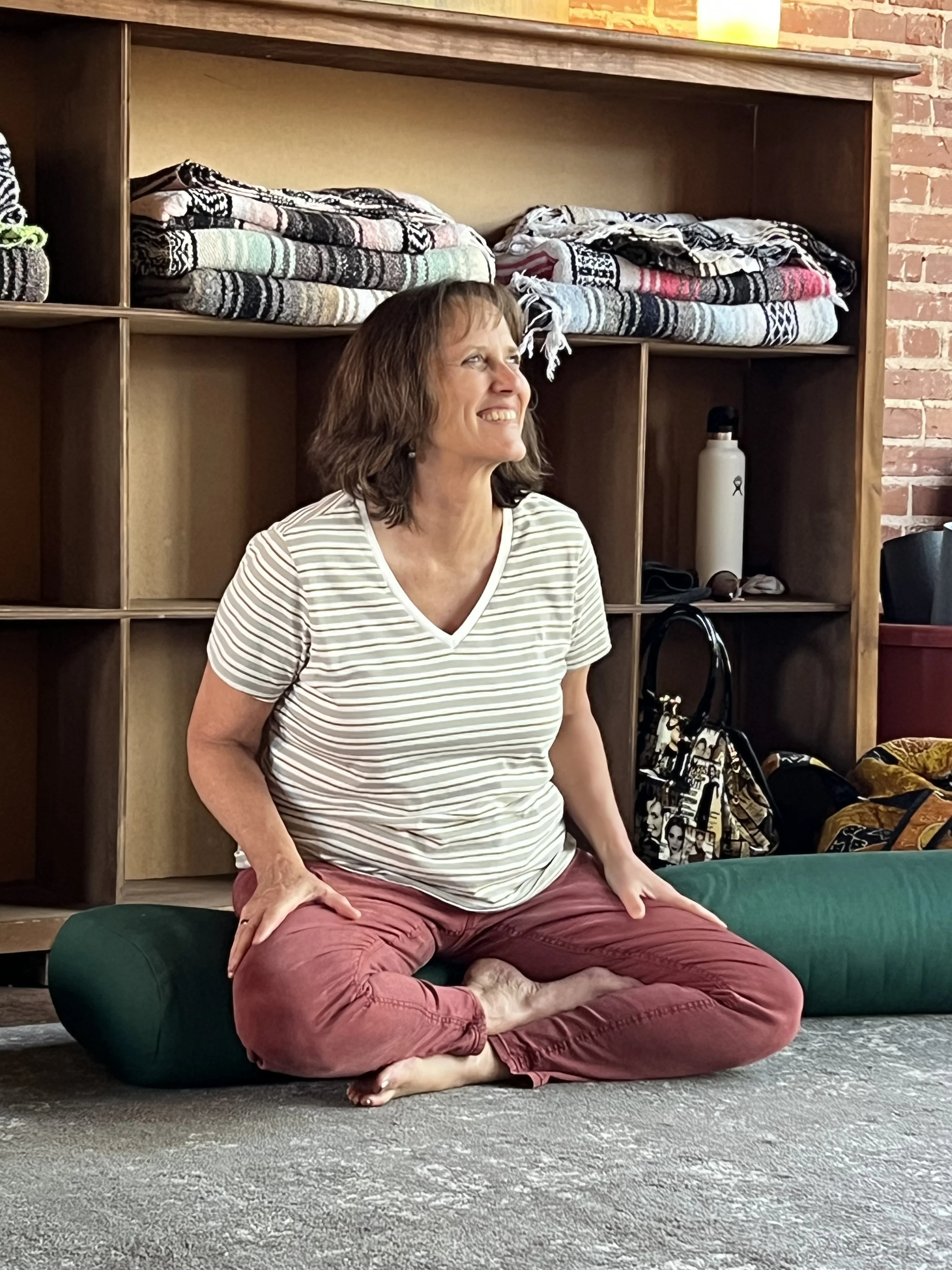 A woman is sitting on the floor in a meditative pose on top of a green cushion. She is smiling and looking to her left, wearing a striped t-shirt and red pants. Behind her, there is a wooden shelf with folded blankets and other items, a brick wall, a