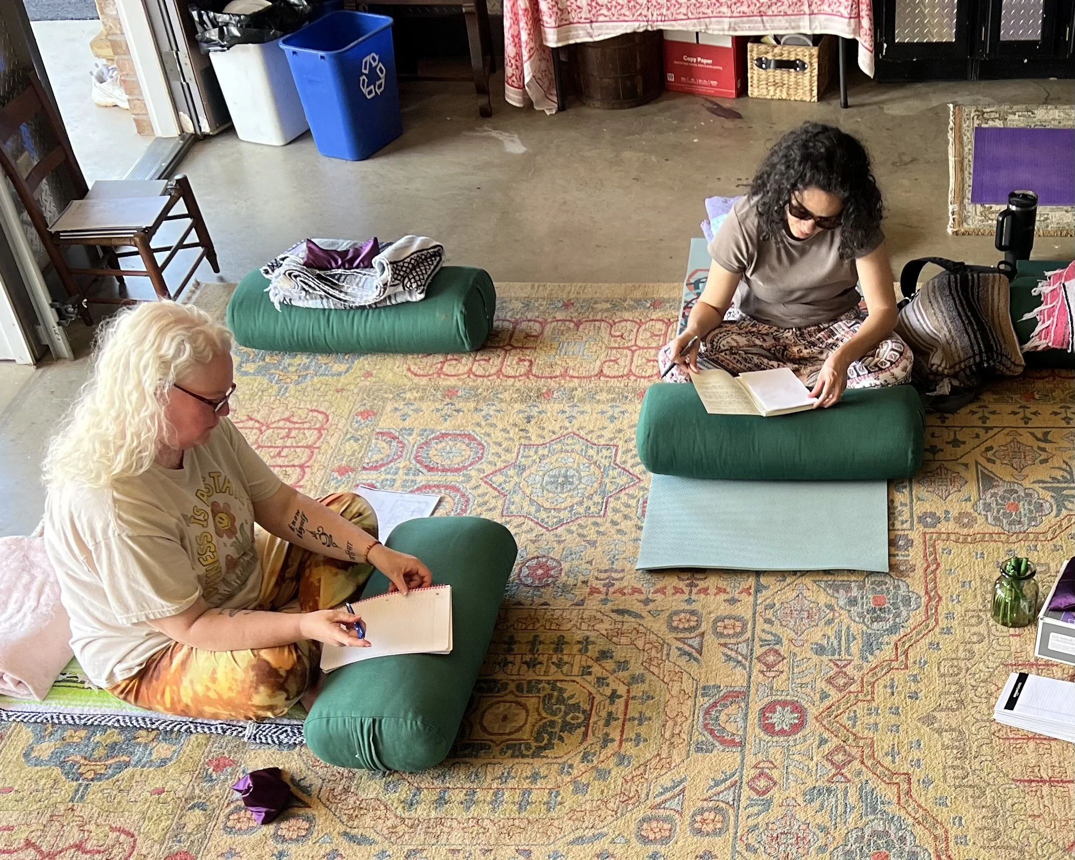 Two women sitting on the floor in a room with oriental-style carpet, engaged in reading or writing activity. One woman with curly dark hair wearing glasses and a gray shirt, the other with long blonde curly hair wearing glasses and a beige t-shirt. There are various items like a tablet, notebooks, and a water bottle around them.