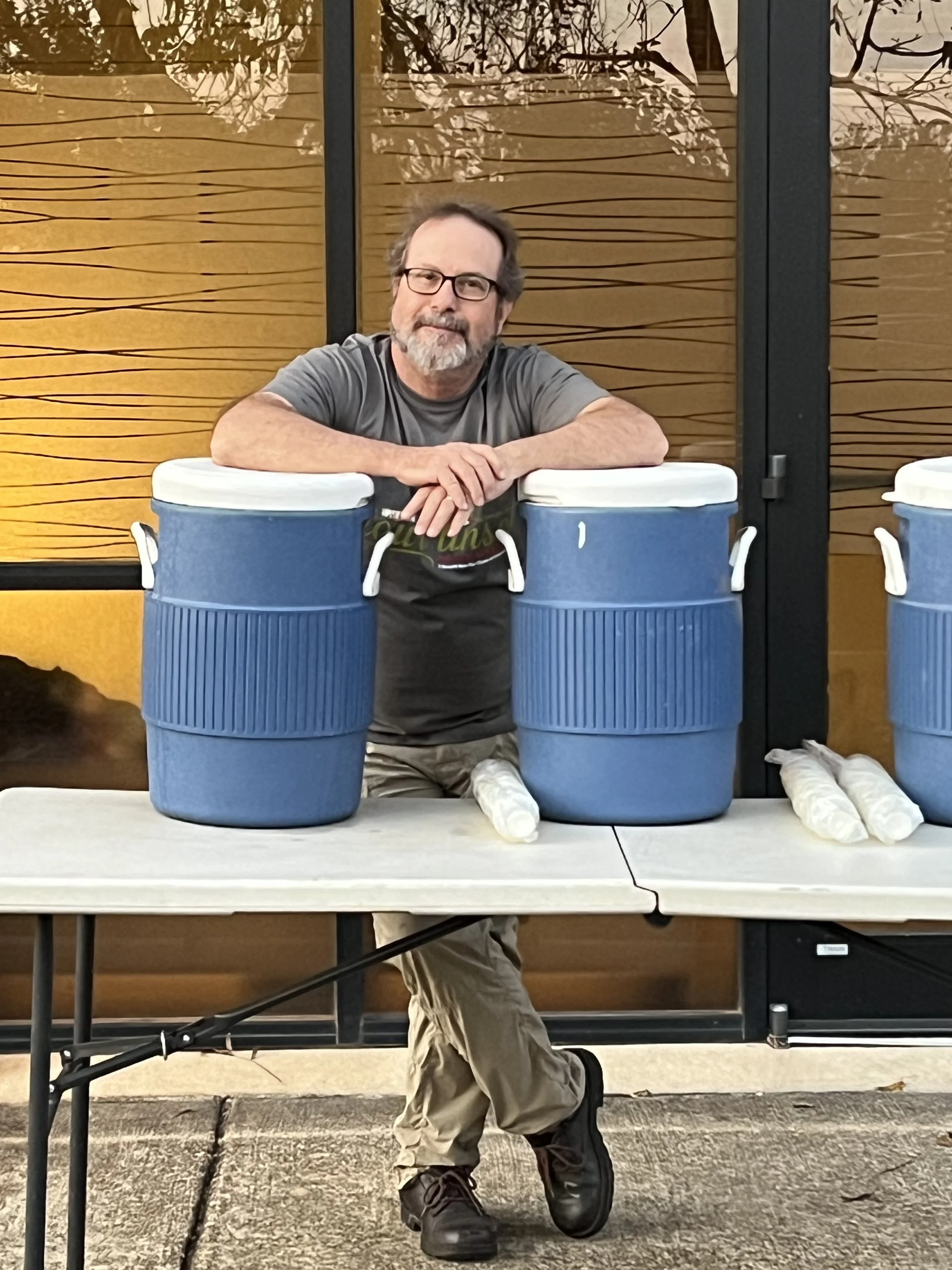 A man with glasses, a beard, and gray hair rests his arms on two large blue coolers on a table. He appears to be at an event, possibly involving food or drinks.