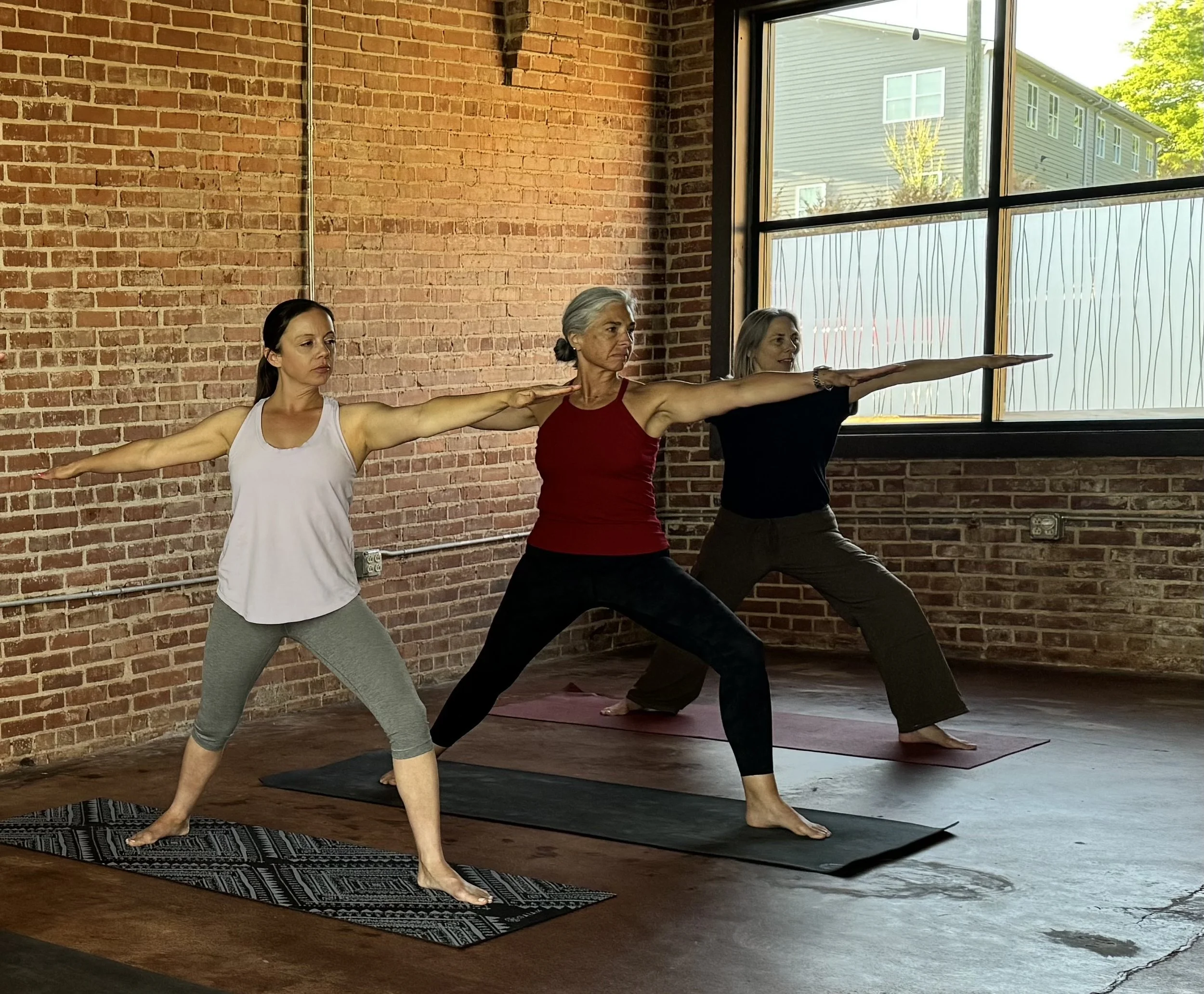 Three women practicing yoga indoors with brick walls and large windows.