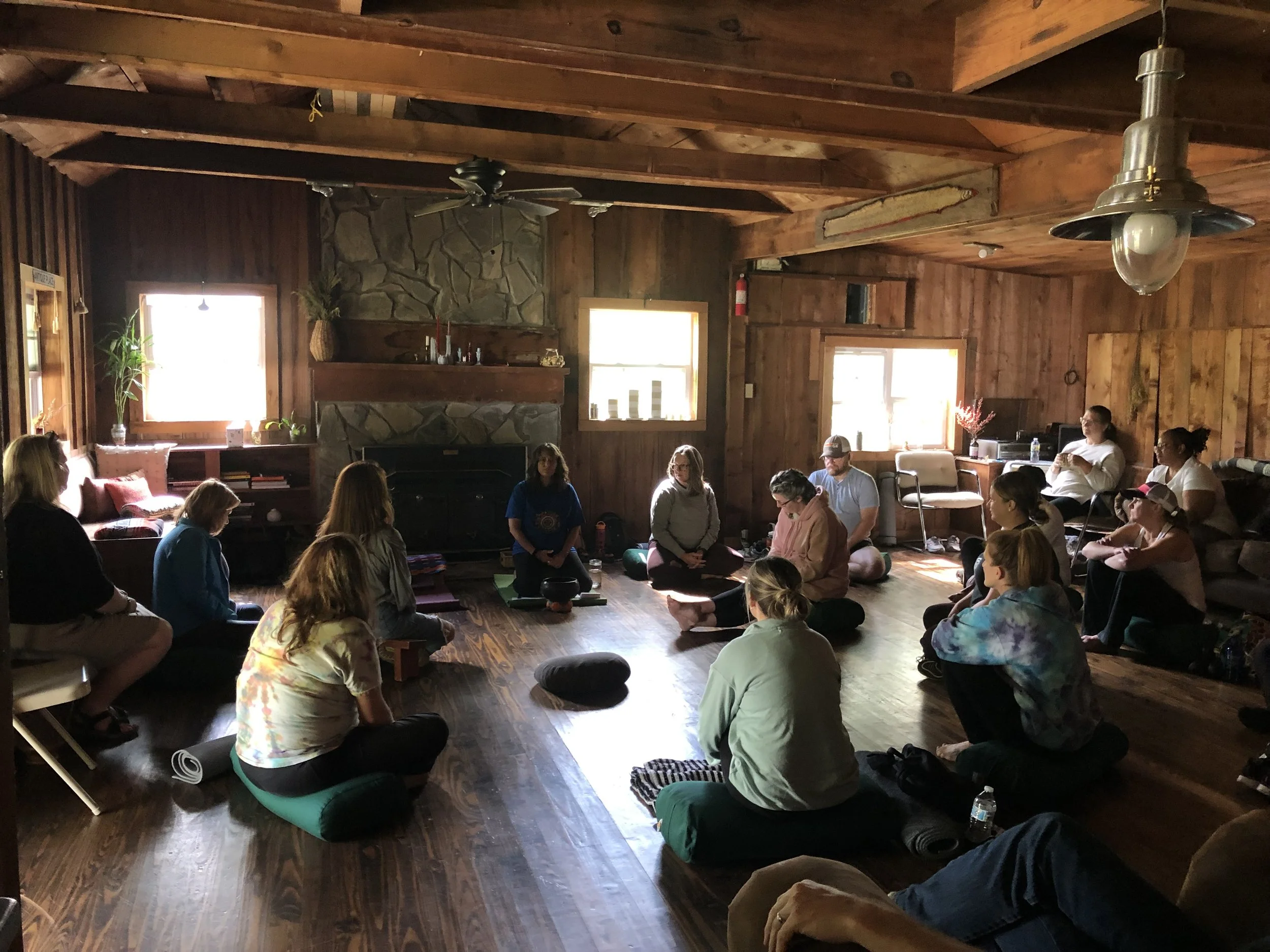 People sitting cross-legged on yoga mats in a cozy, wood-paneled room with a stone fireplace, engaged in a meditation or yoga session during daytime.