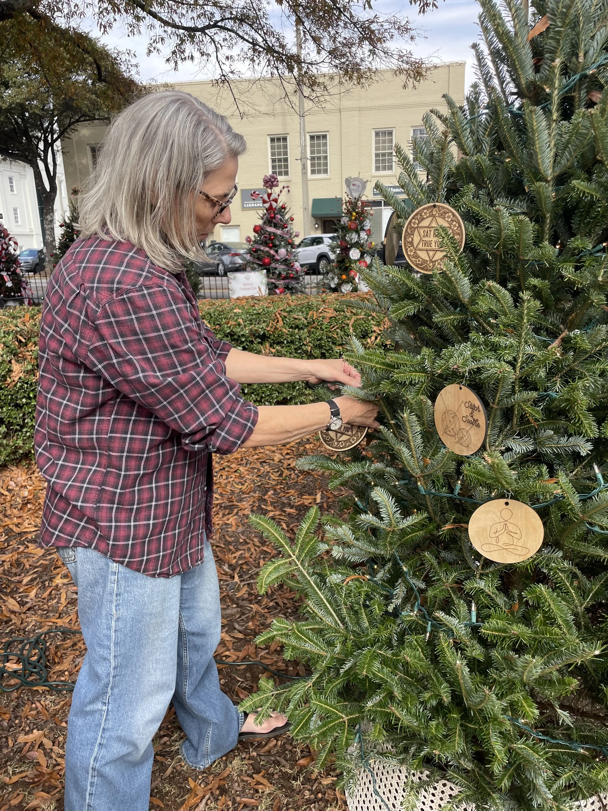 A woman with gray hair and glasses, wearing a red plaid shirt, stands outdoors near a decorated Christmas tree surrounded by fallen leaves, with buildings and parked cars in the background.