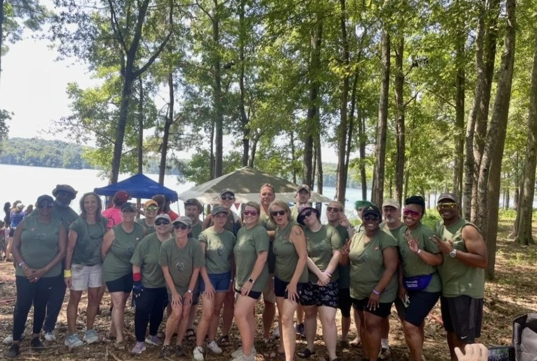 Group of people in matching green shirts outdoors near a lake, with trees and tents in the background.