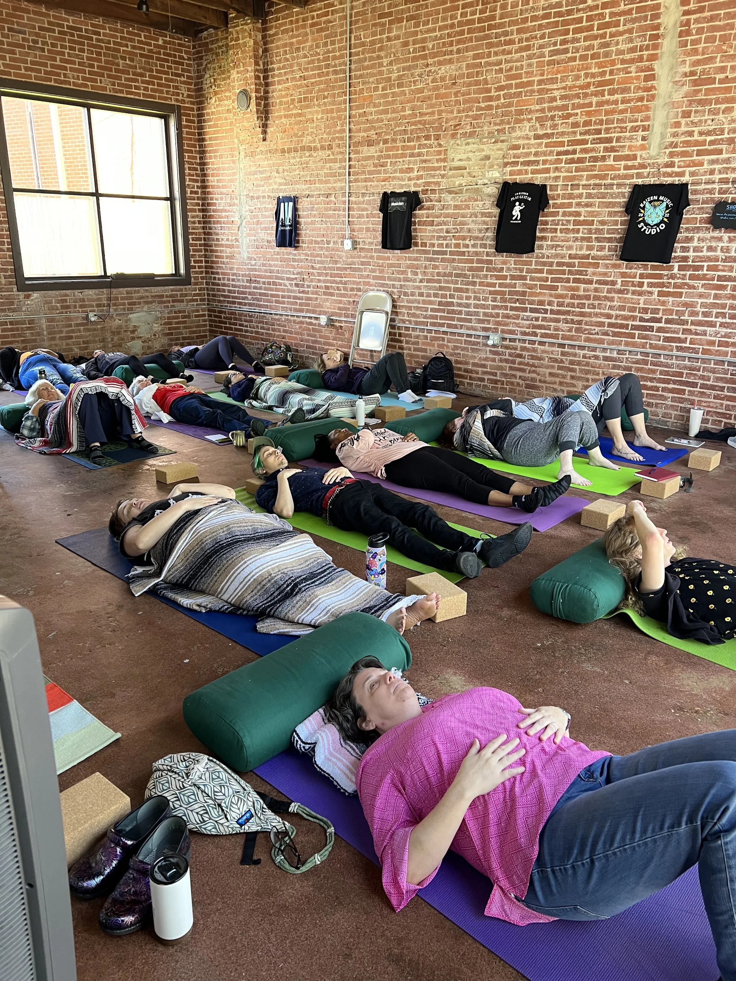 People meditation indoors on colorful mats, lying on their backs with eyes closed, on a wooden floor with brick walls and a window.
