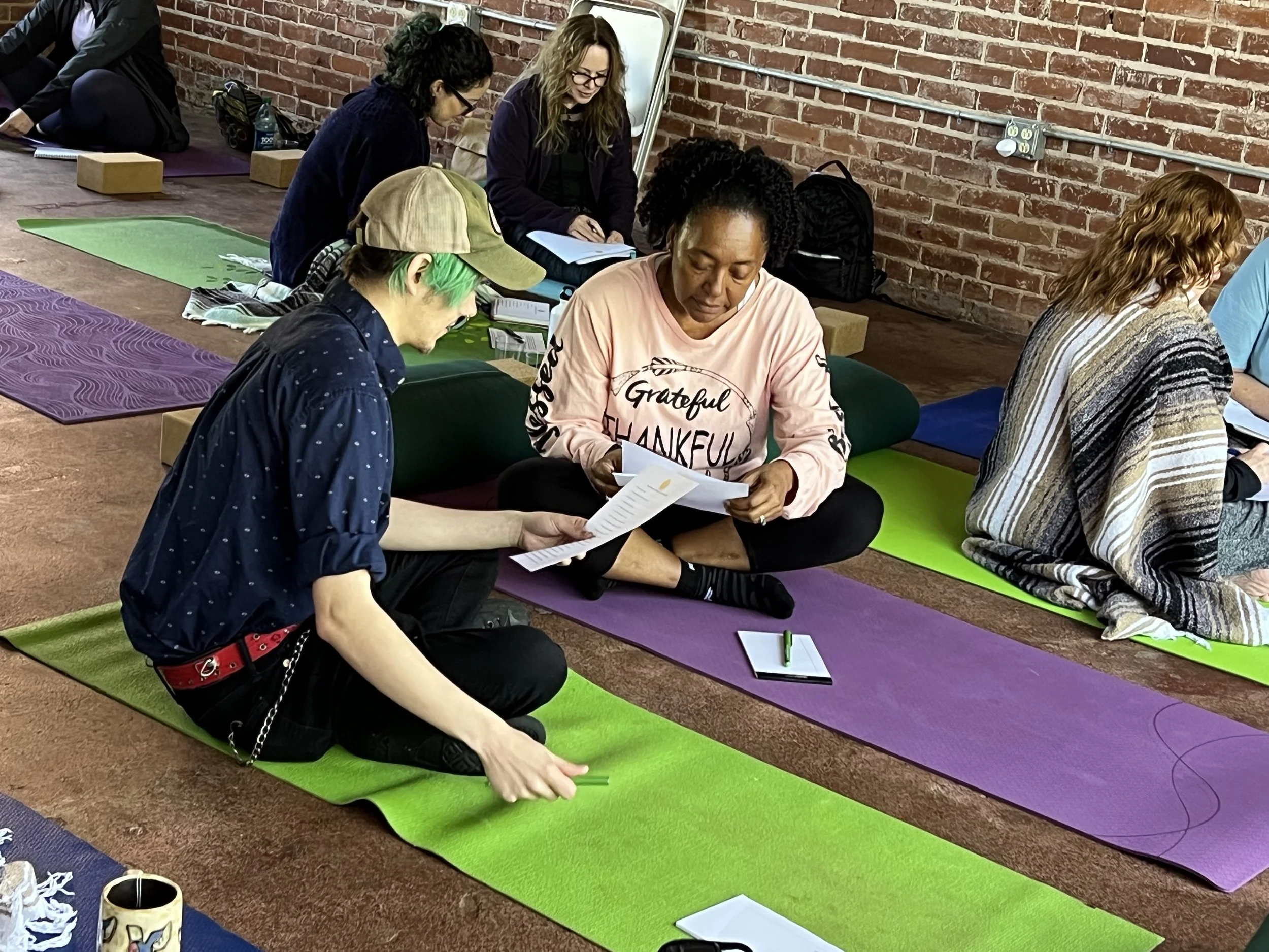 People participating in a class, sitting on colorful yoga mats on the floor against a brick wall, engaging with papers and discussing.