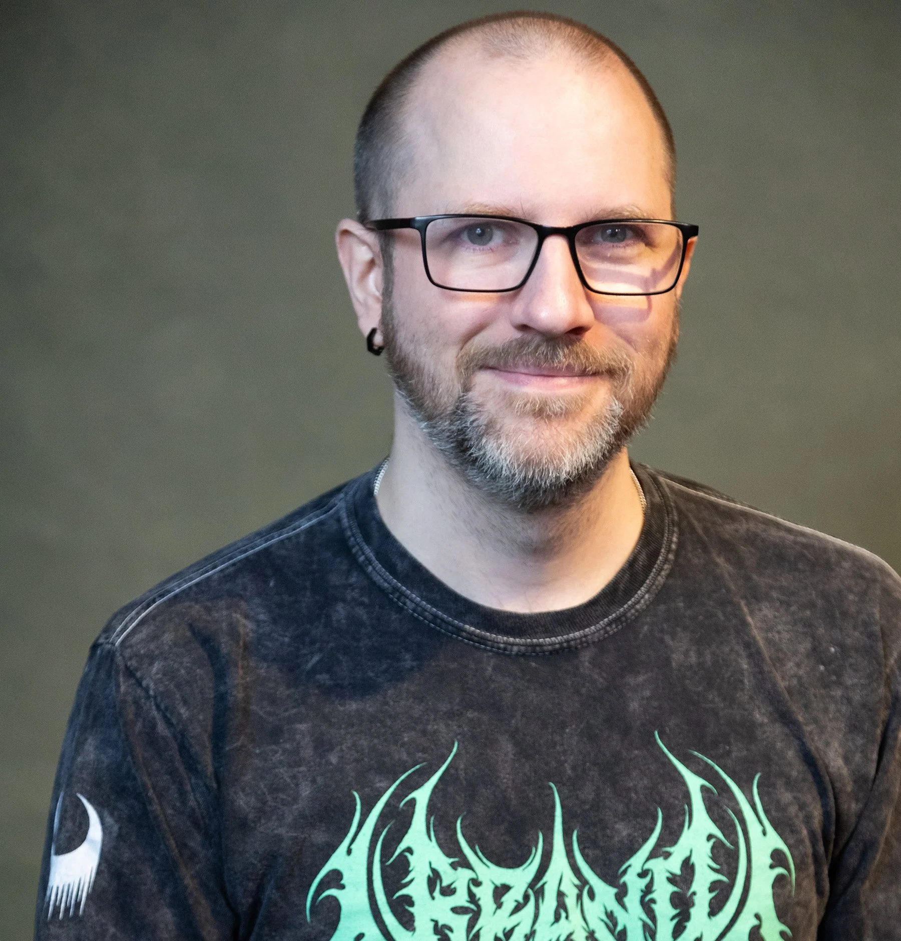 A man with glasses, facial hair, and a shaved head, wearing a black T-shirt with green graphic text, smiling against a plain background.