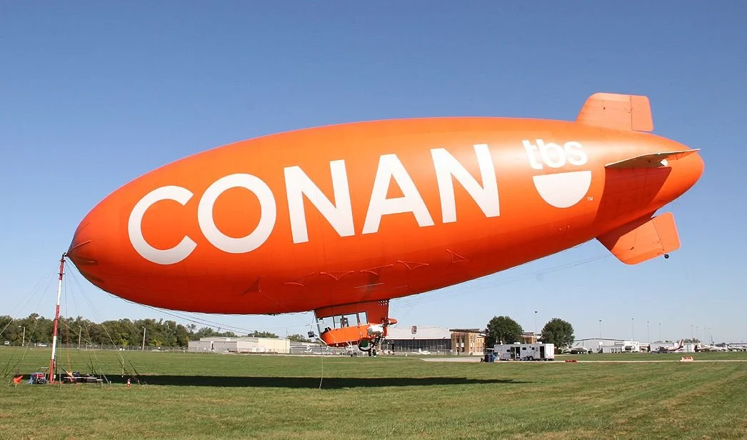 An orange blimp with the words 'CONAN TBS' written on it, floating low over a grassy field.