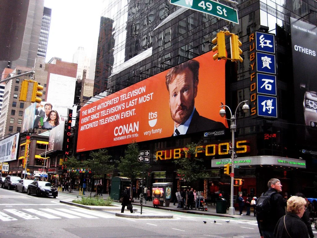 Billboard advertising the television show Conan on a city street corner in Times Square, New York, with people walking and cars on the street.