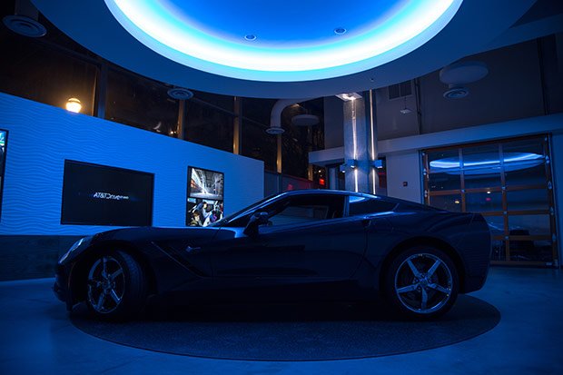 Black sports car displayed indoors with blue lighting and digital screens on the wall in a modern showroom or exhibition space.