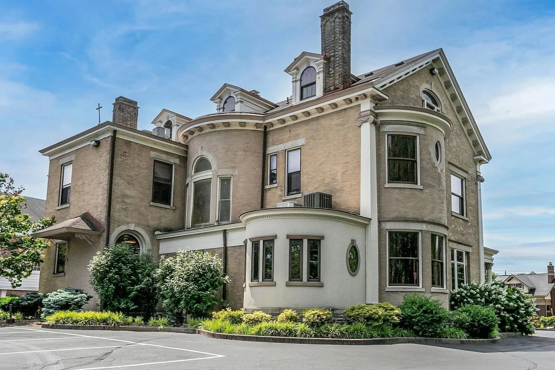 A large historic multi-story house with brick and white stucco exterior, bay windows, and decorative architectural details, surrounded by greenery and a parking lot.