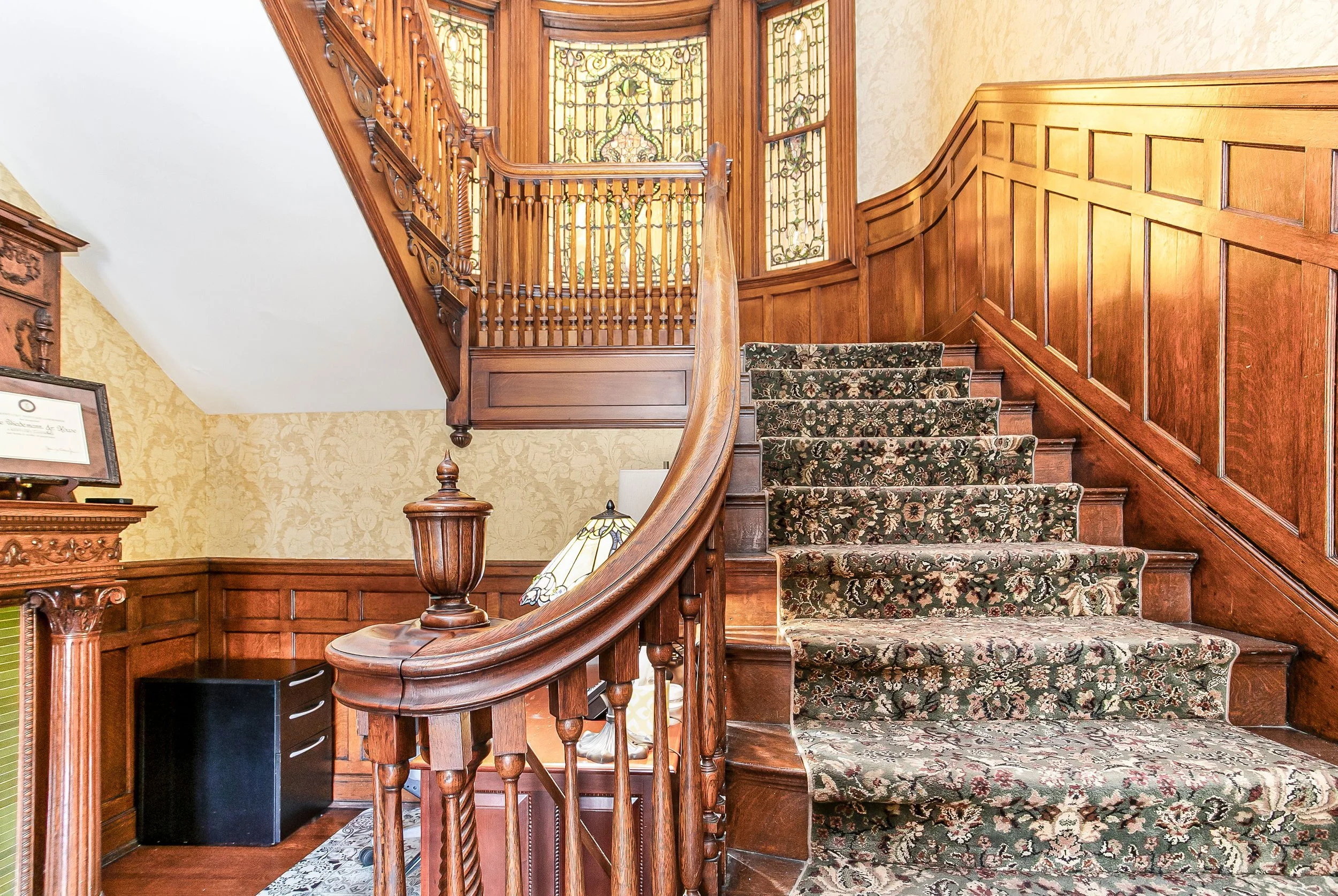 Wood staircase with floral carpet runner, stained glass windows at the top, and wooden paneling on the walls.