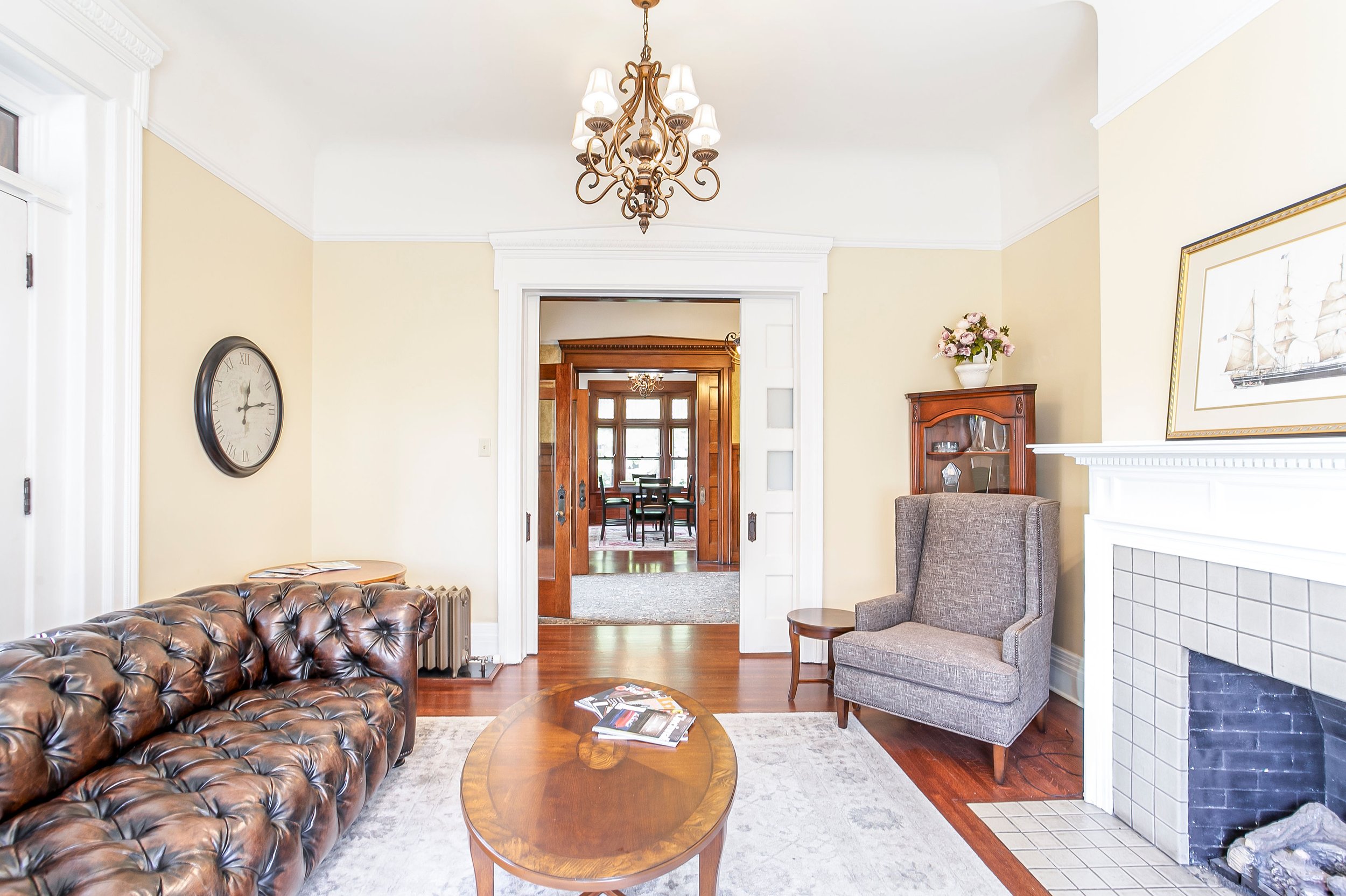 Living room with leather sofa, vintage clock, gray armchair, fireplace, chandelier, and doorway to dining area with large windows.