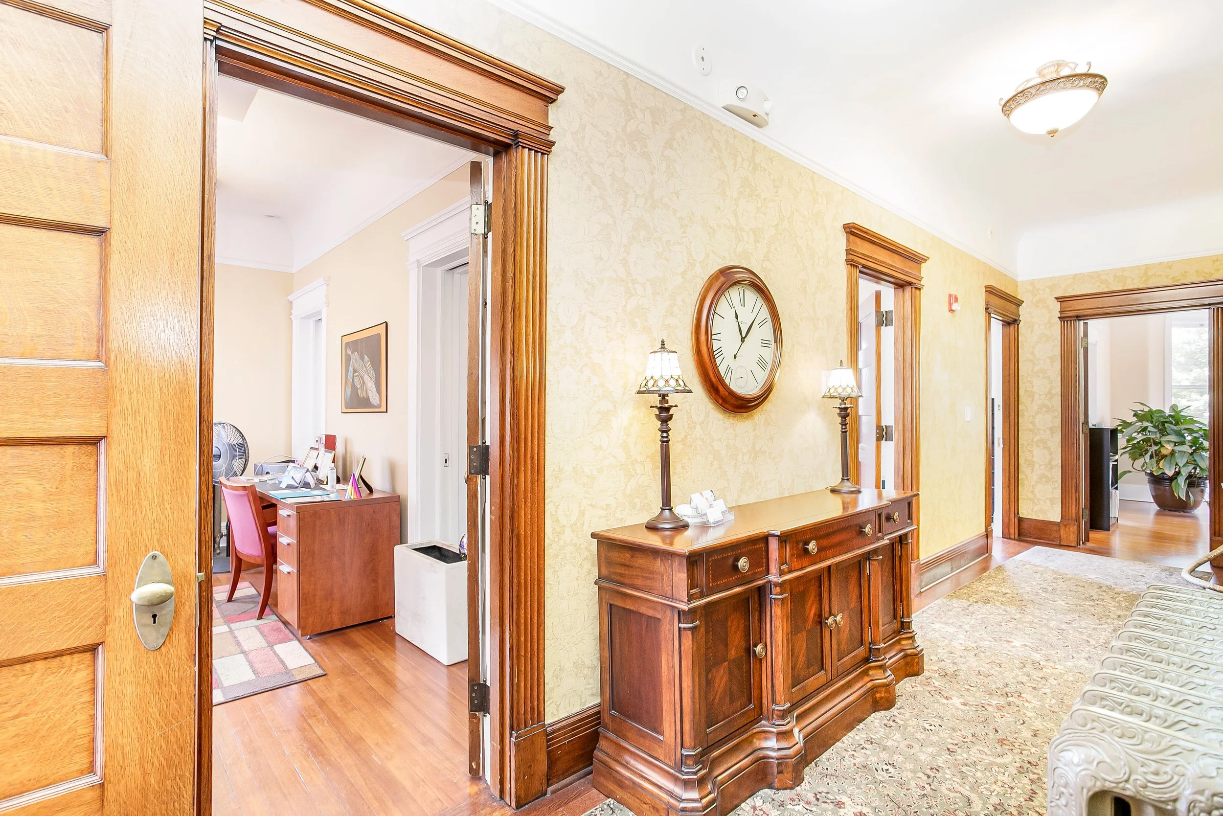 Interior of a house entryway with a wooden console table, two lamps, a large wall clock, an open door leading to a room with a desk and chair, and a hallway with a window and houseplant.