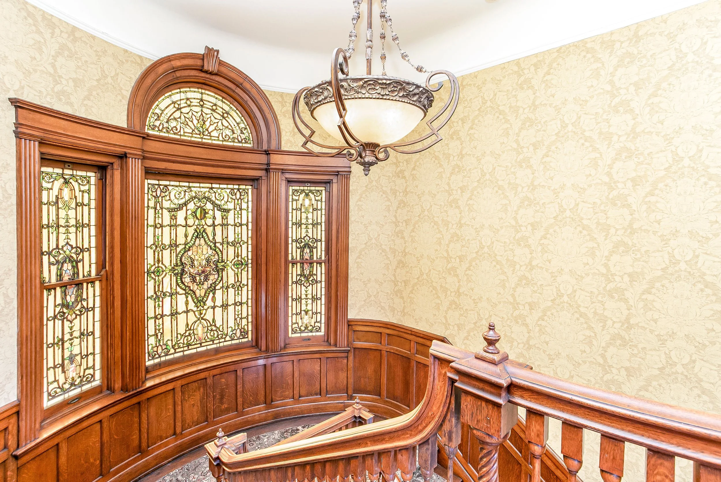 Interior view of a staircase landing with stained glass windows, wooden paneling, and a decorative chandelier hanging from the ceiling.