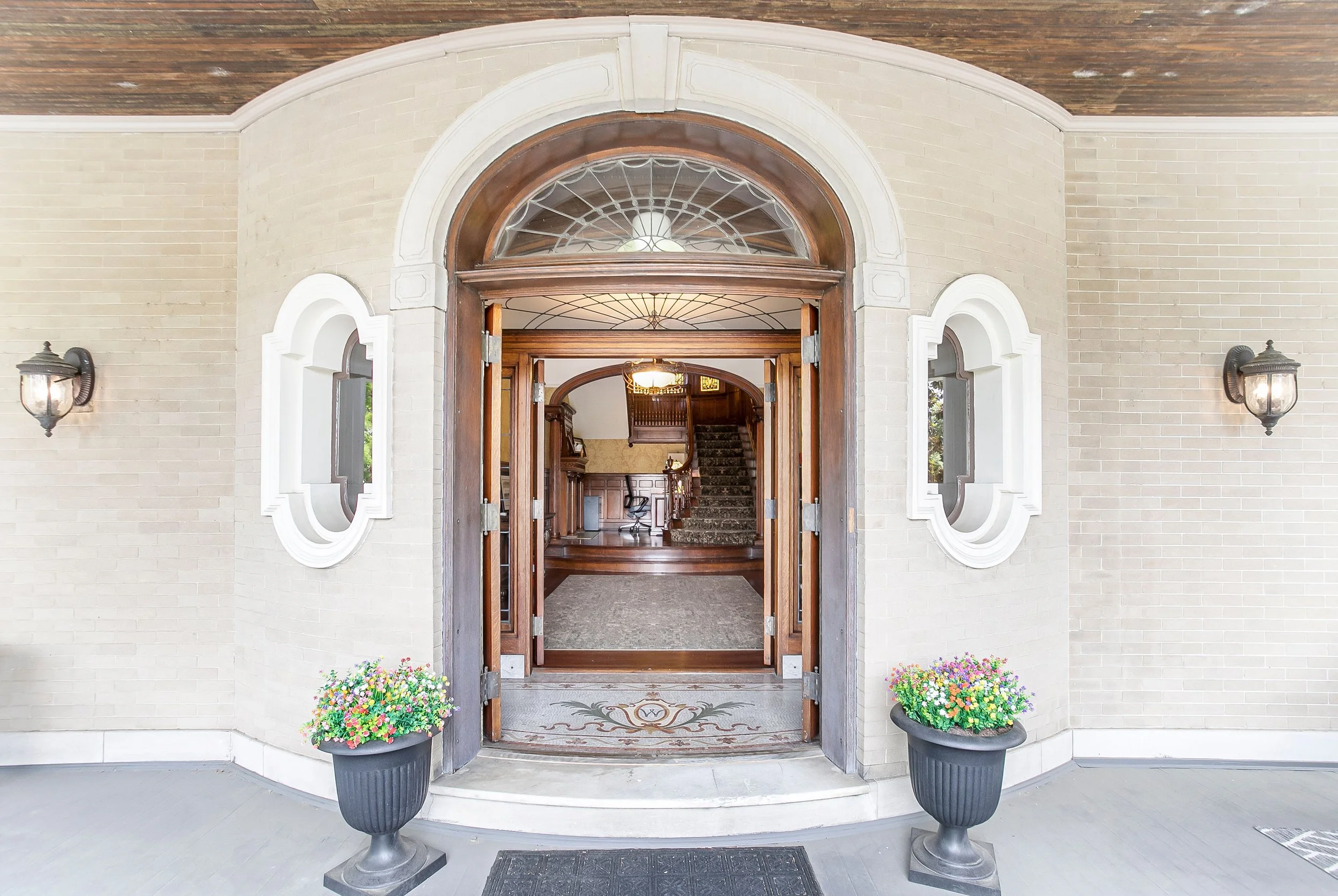 Open wooden front door with glass window panels leading into a house interior, featuring a staircase, a chair, and a chandelier, flanked by white brick walls with outdoor lanterns, and two flower pots with colorful flowers.