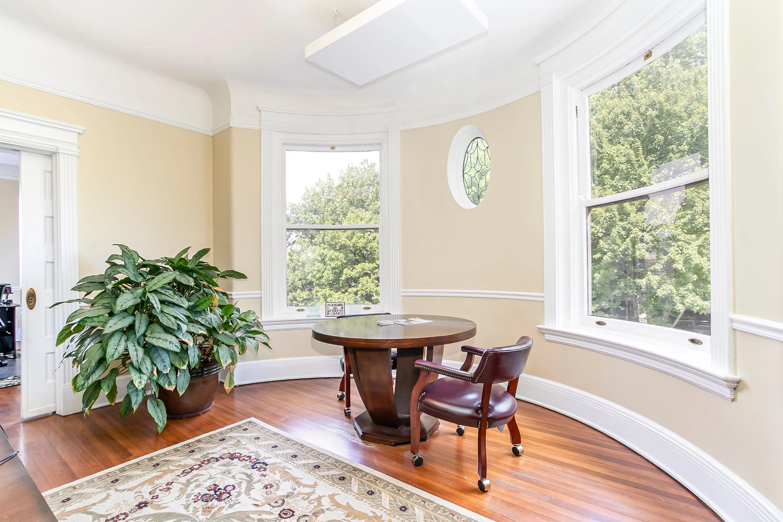 Small office with a round wooden table, a leather office chair, large potted plant, and three large windows with trees outside.
