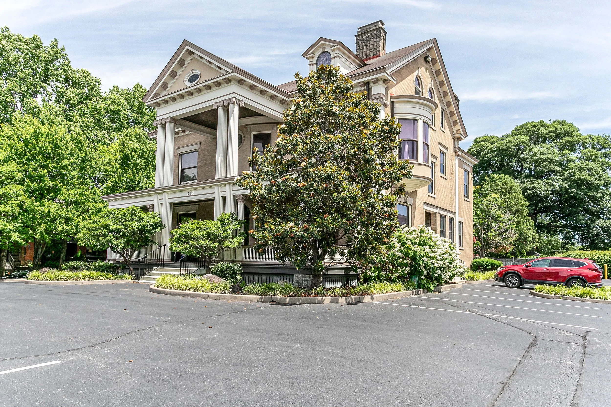 A large, historic house with columns and a porch, surrounded by greenery and trees, with a parking lot and a red car in the foreground.