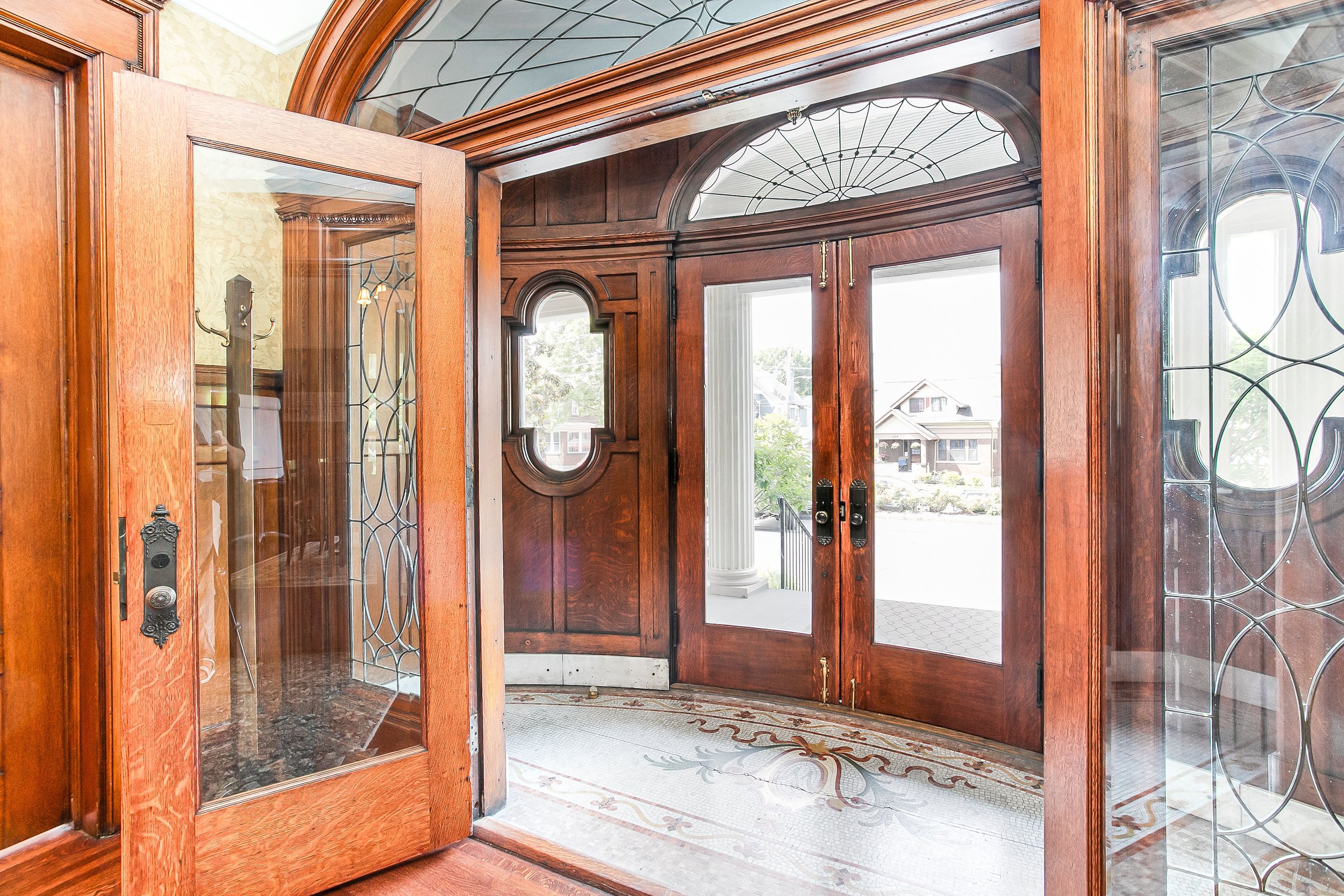 Entryway with wooden doors and glass panels, decorative tile floor, and features outside houses and a porch with columns.
