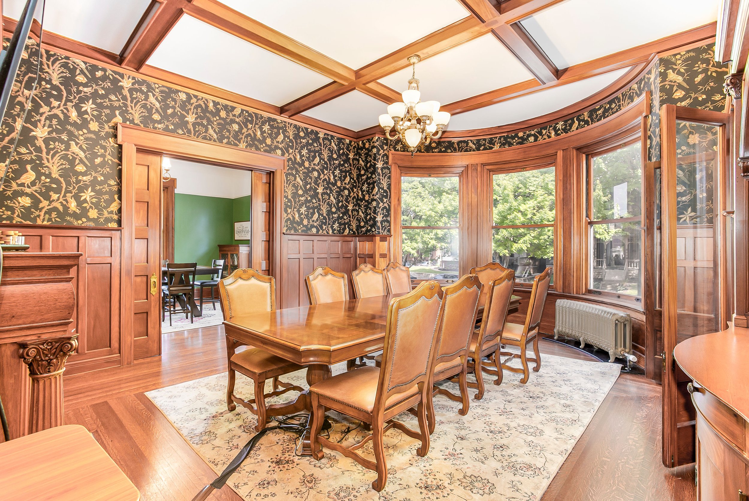 A dining room with wooden wainscoting, floral wallpaper, and large bay windows. There is a long wooden dining table with eight upholstered chairs, a chandelier hanging from the ceiling, and a radiator below the windows. A doorway leads to a room with