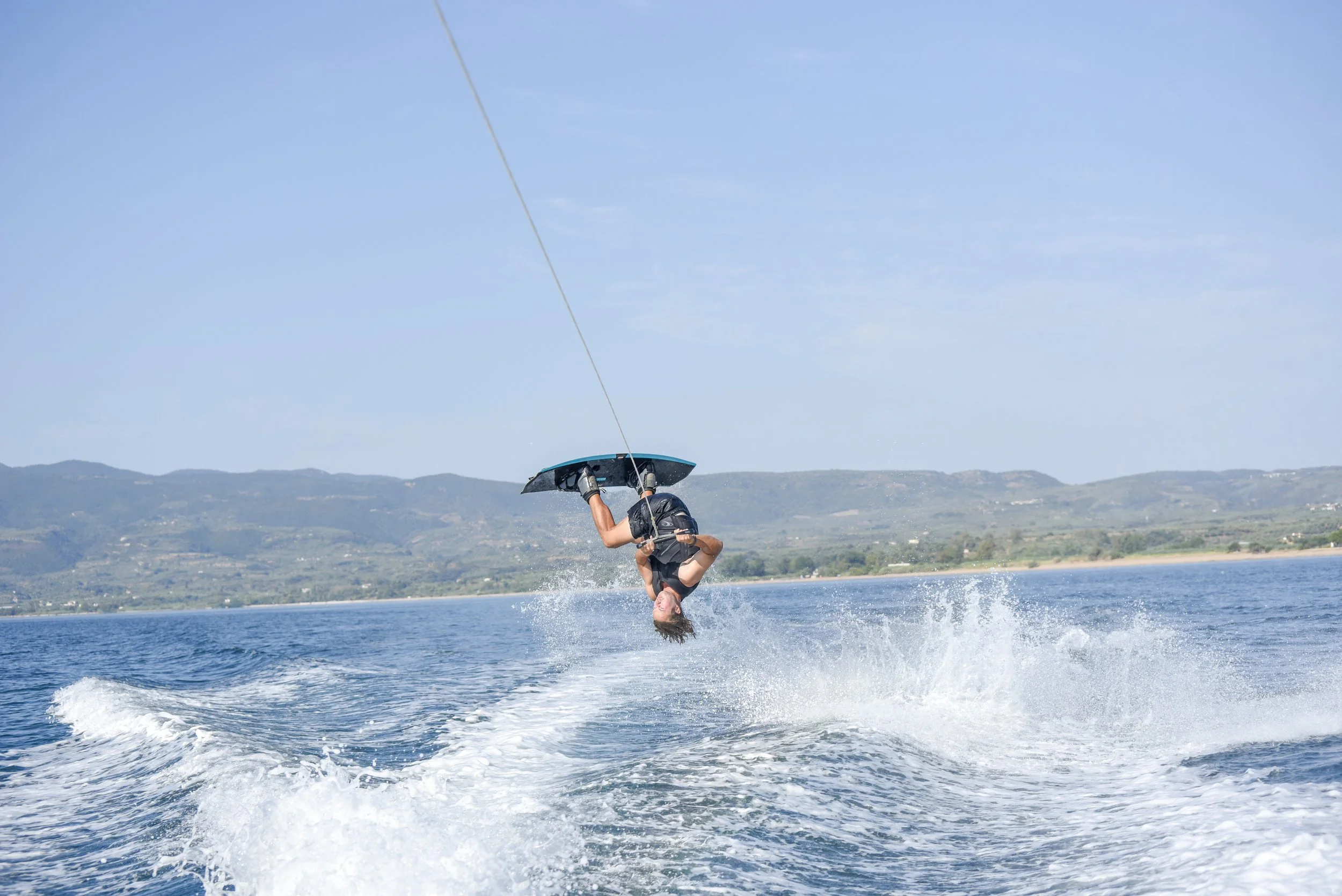 Person performing an acrobatic flip on a wakeboard while being pulled by a boat on a lake, with hills in the background on a clear day.