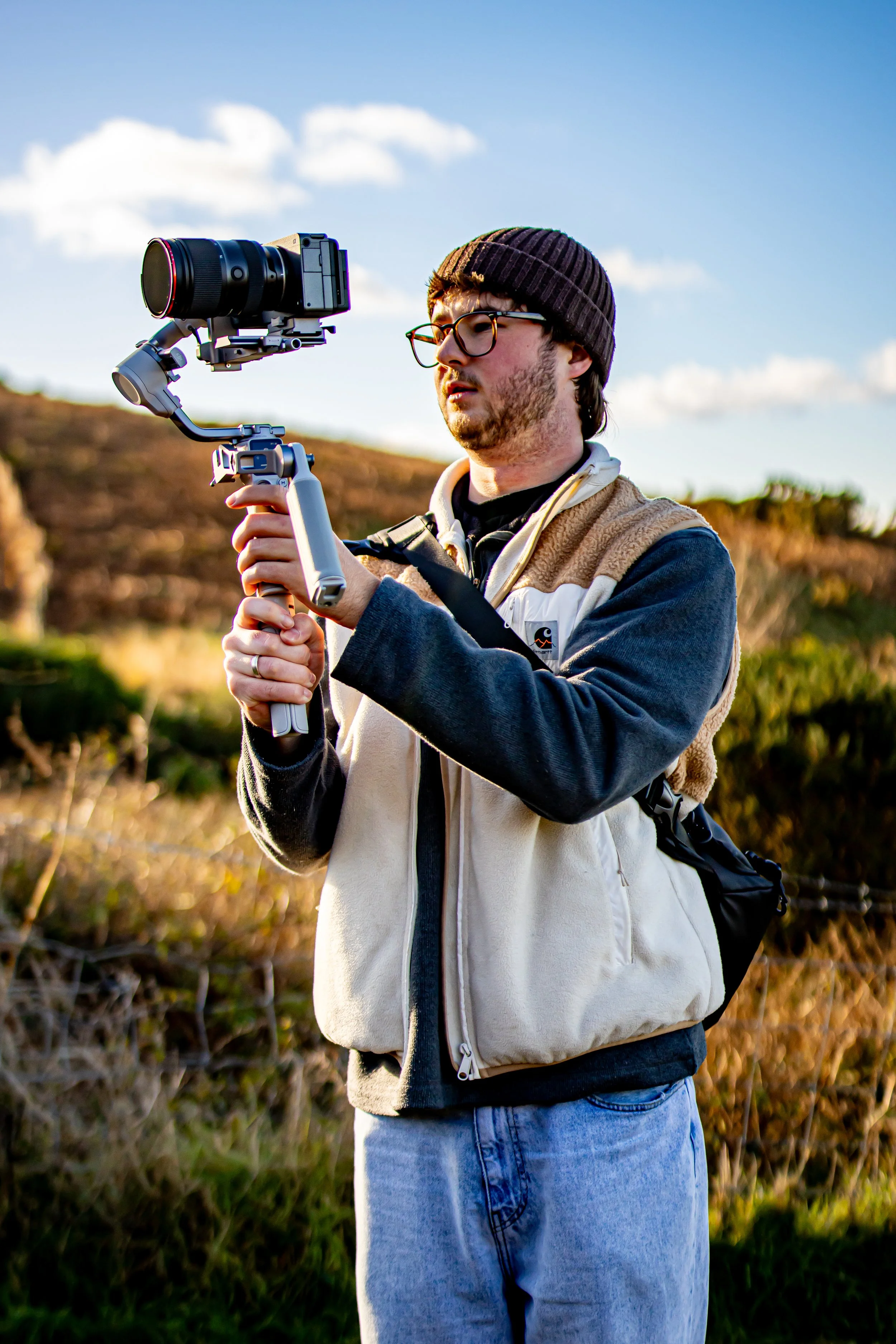 A man outdoors wearing glasses and a beanie, holding a camera stabilizer with a camera attached, filming or taking photos.