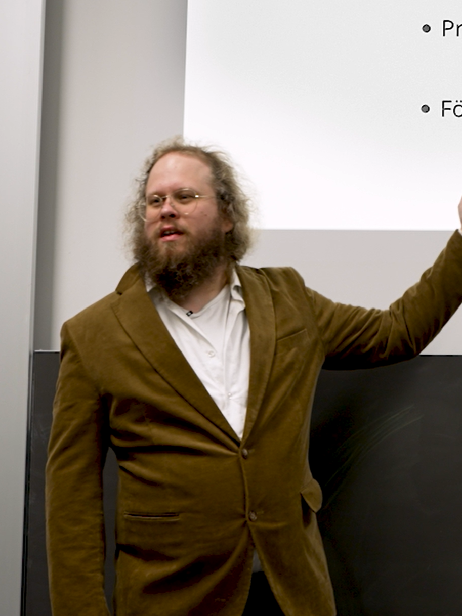 Mann mit Brille, Bart und lockigem Haar, der vor einer Präsentation steht und auf eine Folie zeigt.