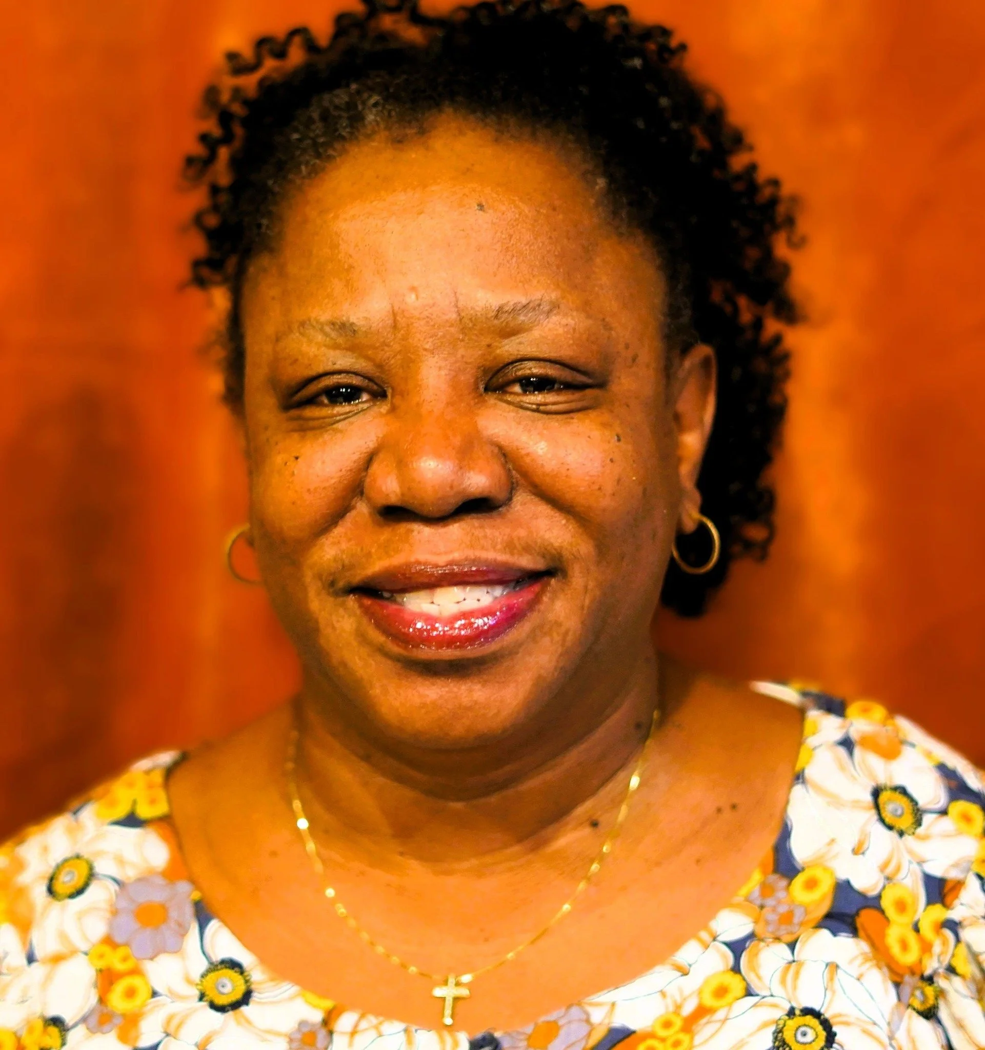 Close-up portrait of a smiling African American woman with curly hair, wearing a floral top, gold hoop earrings, and a gold necklace with a cross pendant, against a warm background.