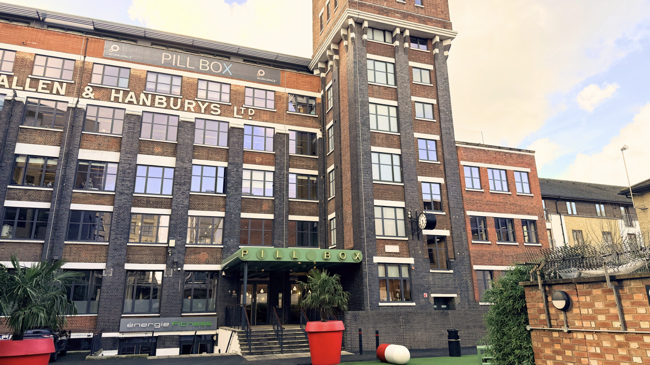 A multi-story brick building with a green sign that reads 'PILLOBOX' above the entrance. The building has several windows and some signs, including 'WORKSPACE' and a clock. There are plants near the entrance and some decorative objects around the front.