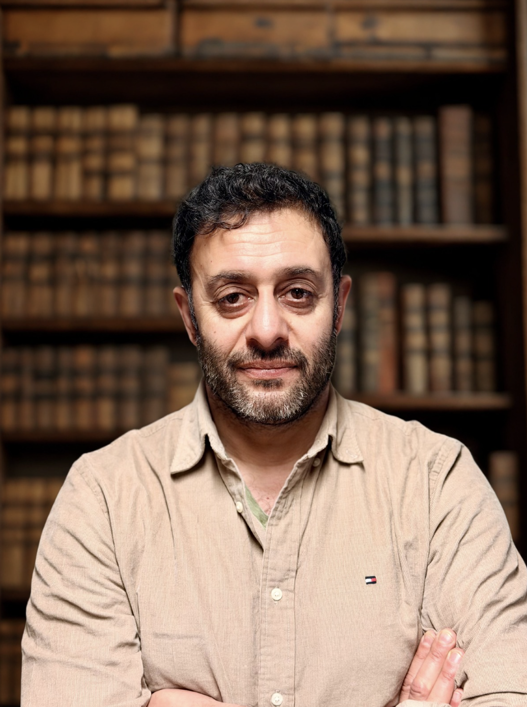 A man with dark hair, a beard, and wearing a beige button-up shirt standing in front of a wooden bookshelf filled with old books.