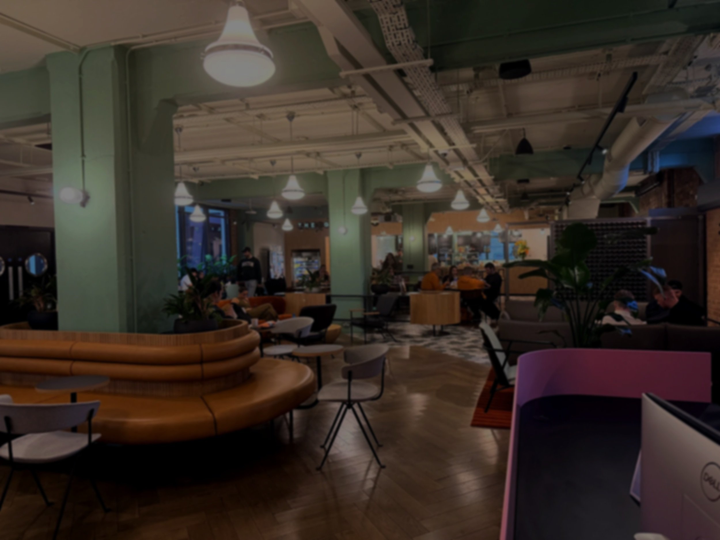 A modern indoor space, possibly a café or lounge, with green pillars, various seating options, and several people sitting and talking. The ceiling has exposed pipes and ductwork, with hanging light fixtures.