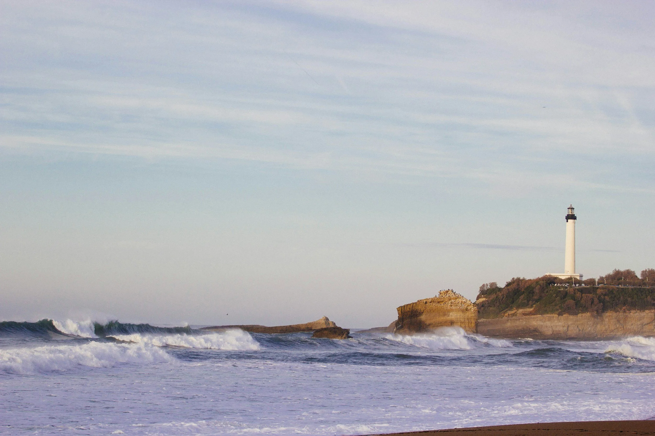 Ocean waves crashing against the shore with a lighthouse on a hill in the background.