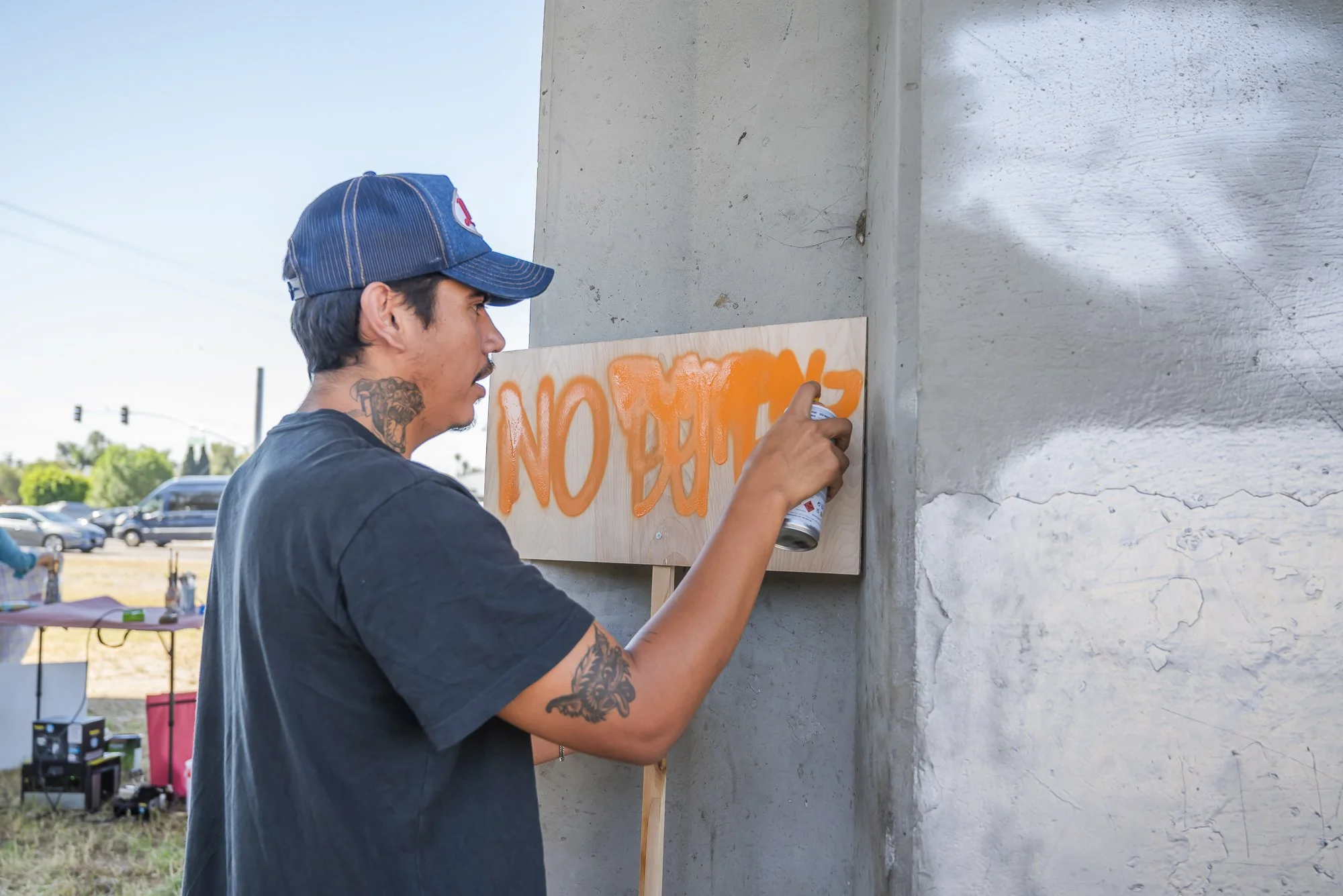 A man with tattoos on his arm and neck, wearing a blue baseball cap and black T-shirt, spray paints orange letters on a wooden sign attached to a grey wall outdoors during daytime.