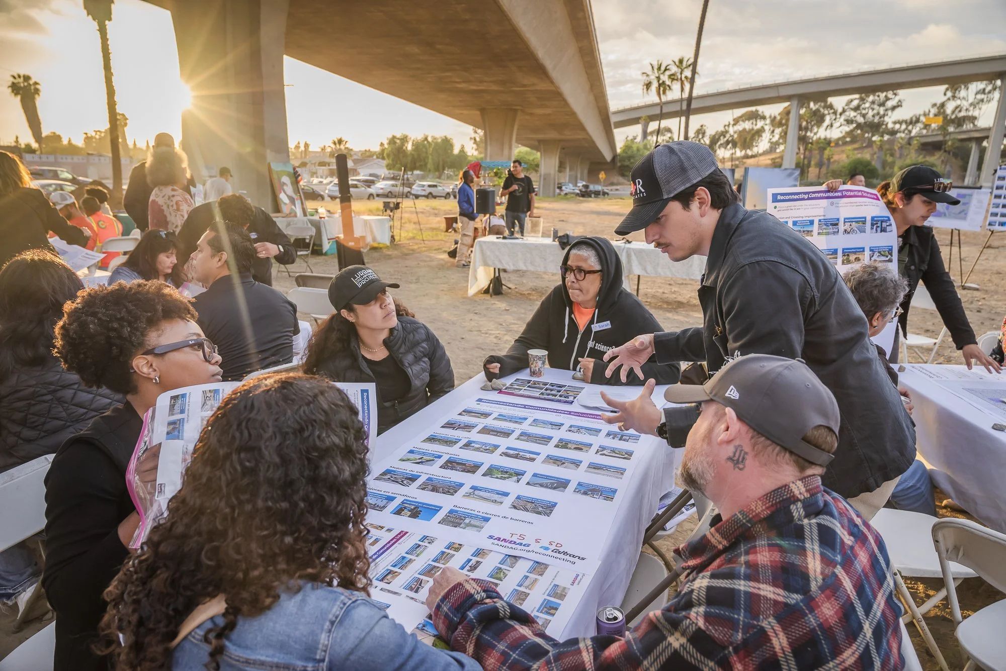 A group of people gathered outdoors at a community event under a bridge during sunset, engaging with informational posters on a long table.