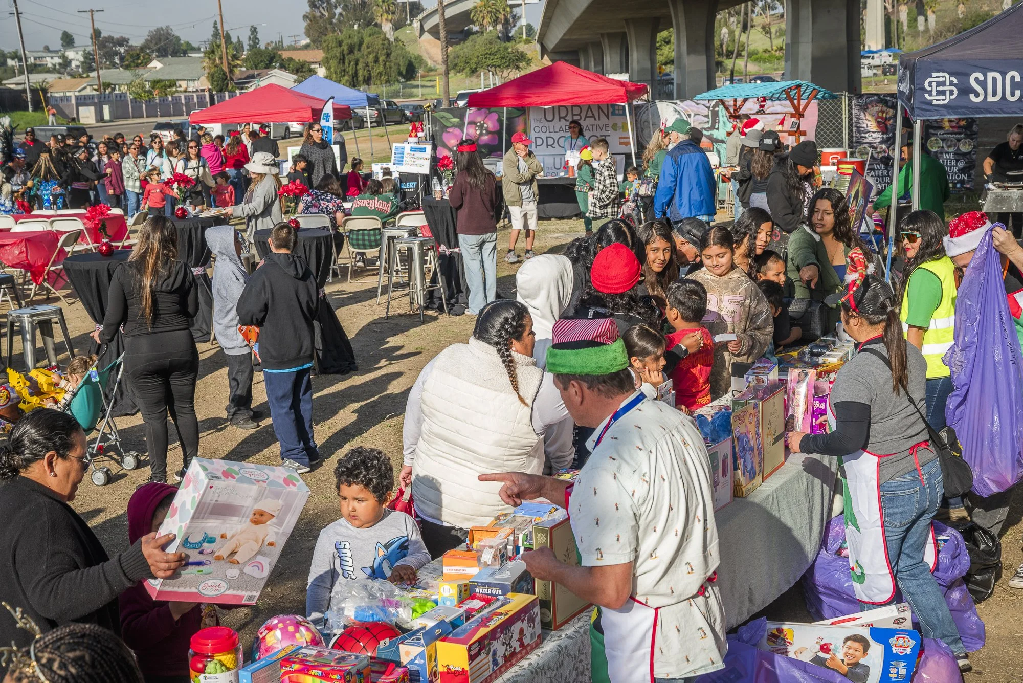 A busy outdoor holiday event with many people shopping at vendor tables, some wearing Christmas hats. There are colorful tents and a backdrop of houses, trees, and an overpass. The scene includes children and adults browsing and socializing.