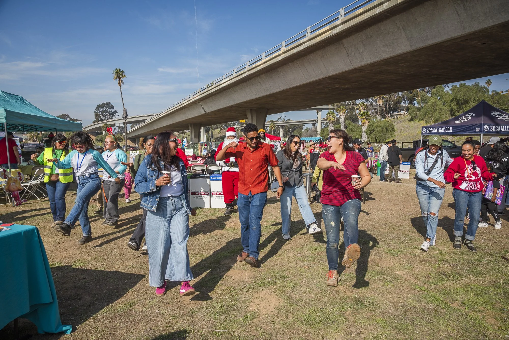 A group of people, some wearing festive attire like Santa hats, enjoying an outdoor event under a bridge with tents and booths around, during a sunny day.