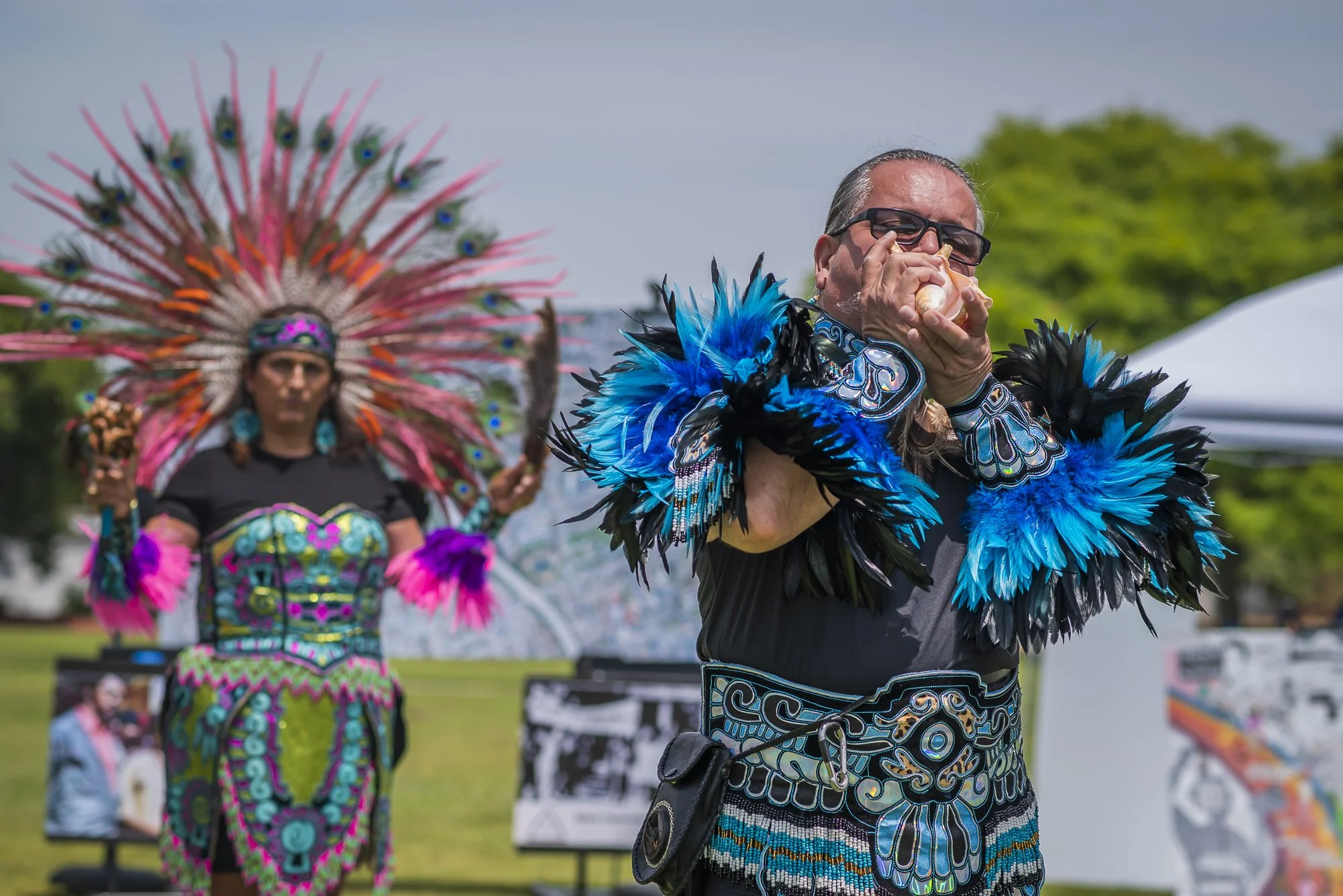 A man dressed in a black shirt with blue and silver feathered tribal costume drinking from a shell during a cultural celebration outdoors, with another person in colorful traditional attire and a headdress in the background.