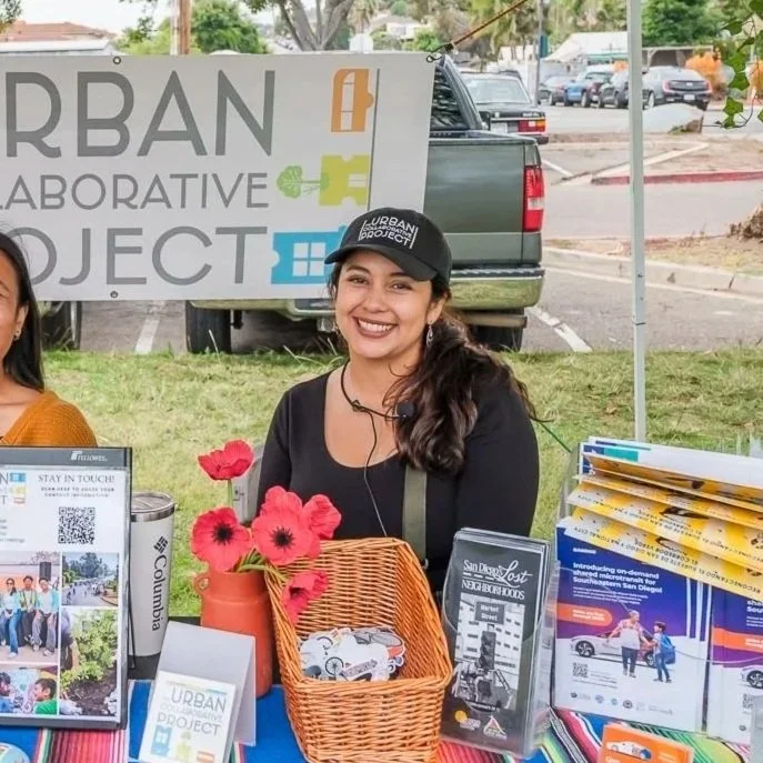 Carmina Paz from the Urban Collaborative project wearing a black cap and black shirt smiles sitting behind a booth at an outdoor event, with informational pamphlets, a basket of flowers, and a sign with the text 'URBAN COLLABORATIVE PROJECT'.