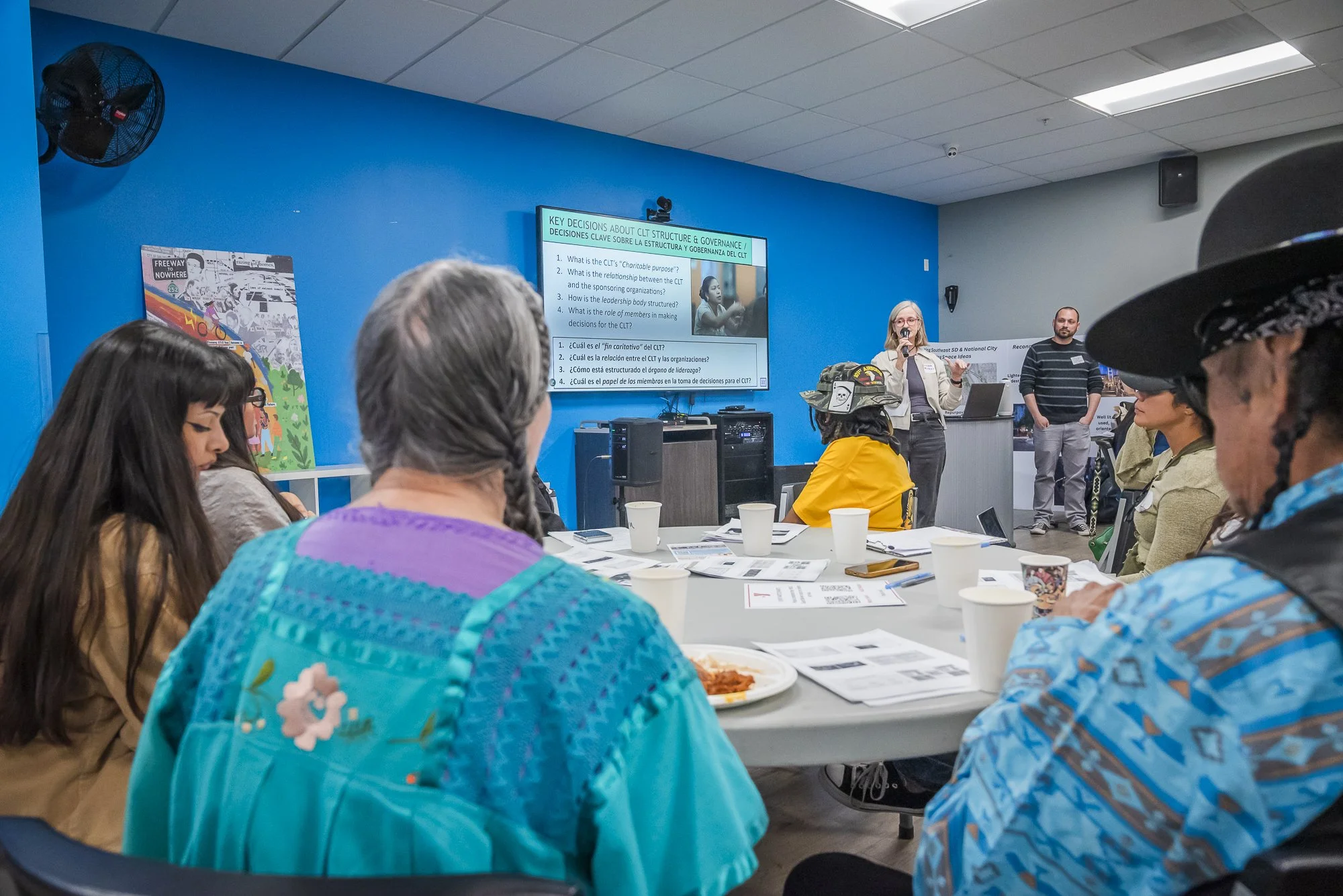 A diverse group of people sitting around a table during a presentation in a conference room. A woman is speaking and gesturing, with a large screen displaying presentation slides behind her. The room has a blue accent wall and colorful artwork, with 