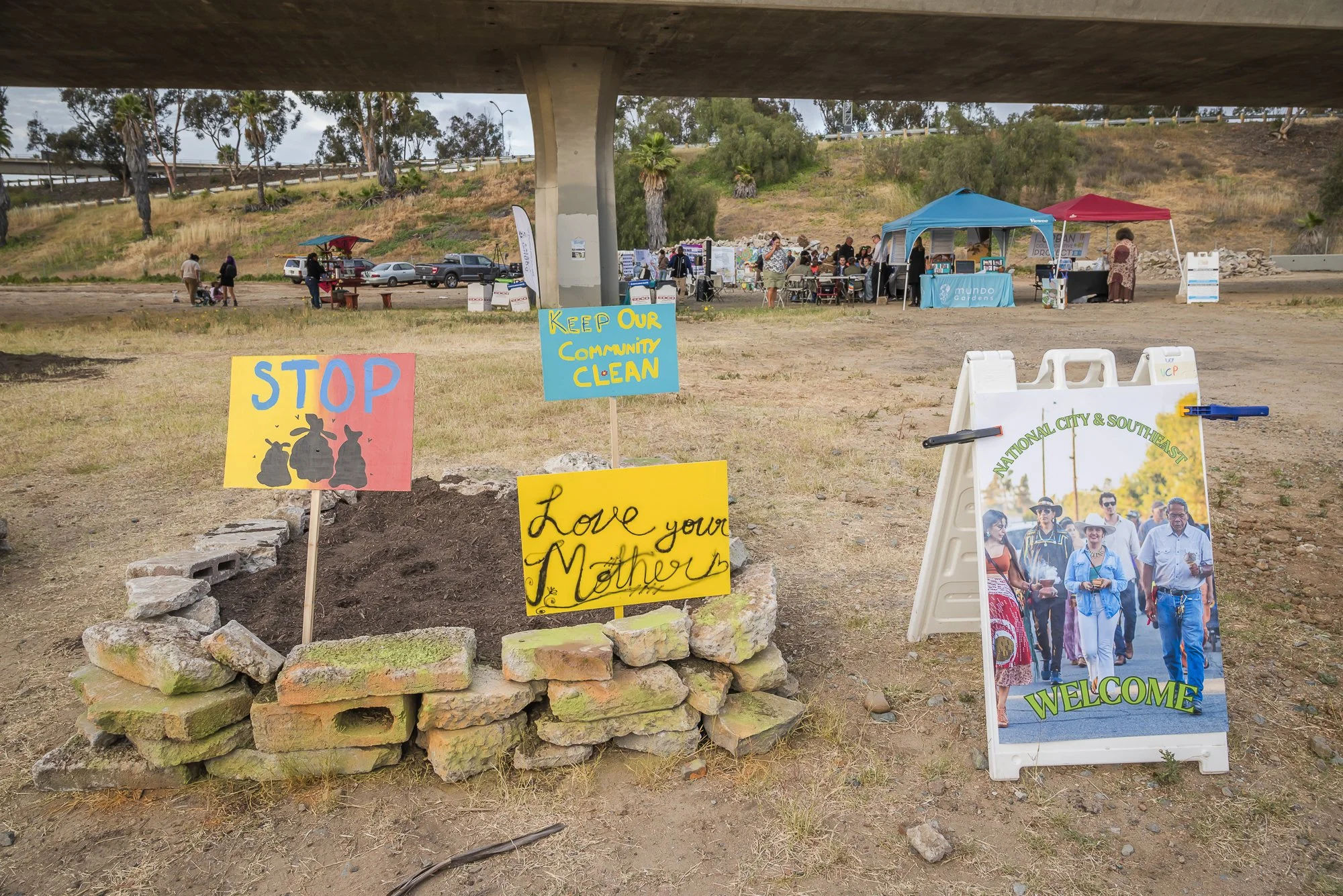 Community event under a bridge with tents and signs. Signs say 'Stop,' 'Keep Our Community Clean,' and 'Love Your Mother.' A group of people is gathered around booths and displays in the background. A photo board on the right welcomes visitors.