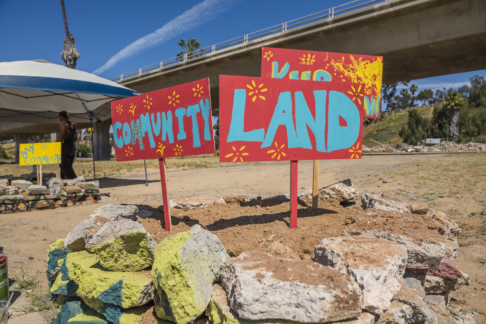 Colorful handmade signs that read "My Community Land" and are decorated with yellow and red flowers, set among painted rocks in an outdoor area with tents, trees, and an overpass in the background.