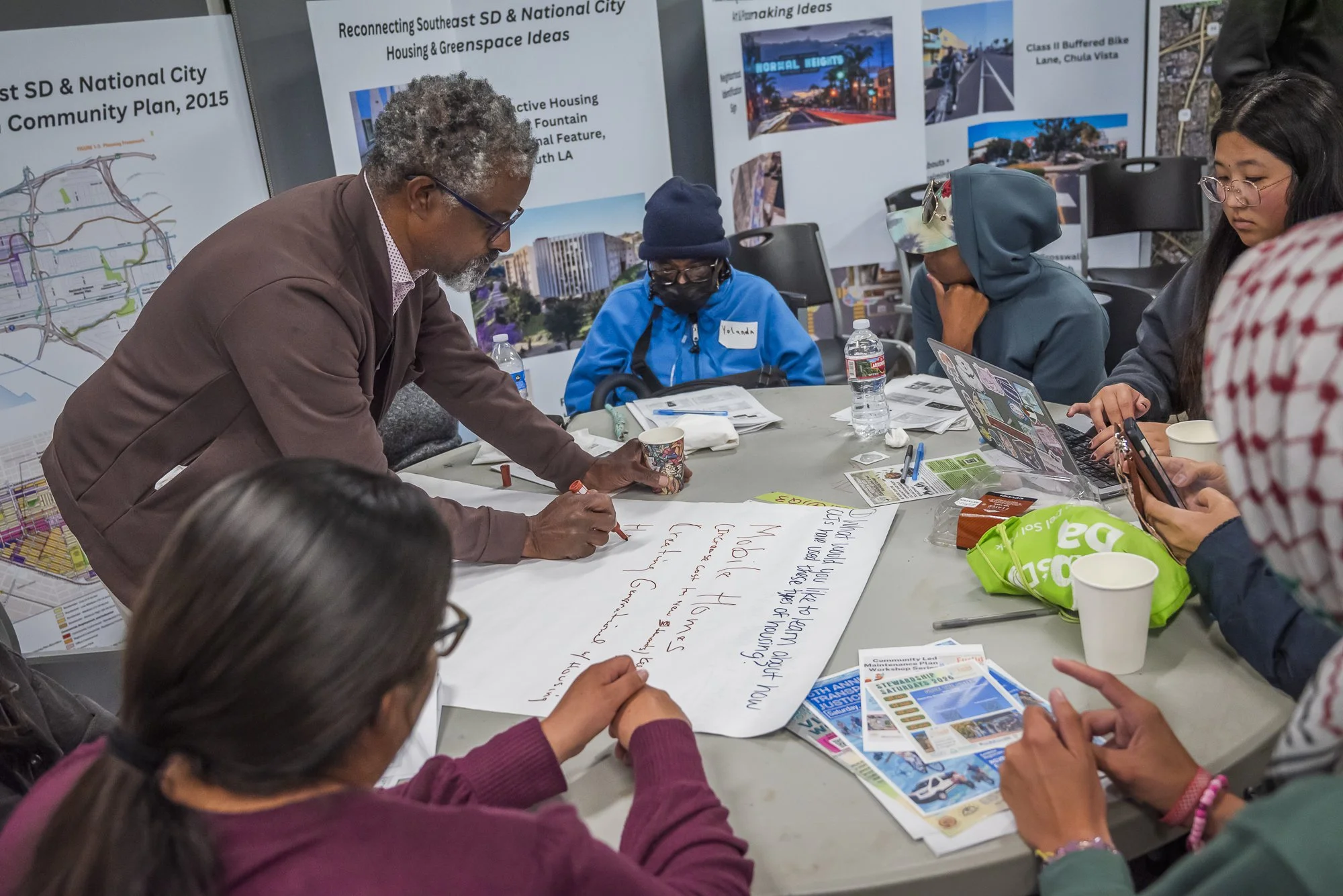 A group of diverse people gathered around a table working on a community planning project. One person is writing on a large sheet of paper, with posters and maps on the wall behind them showing development and housing ideas for Southeast San Diego.