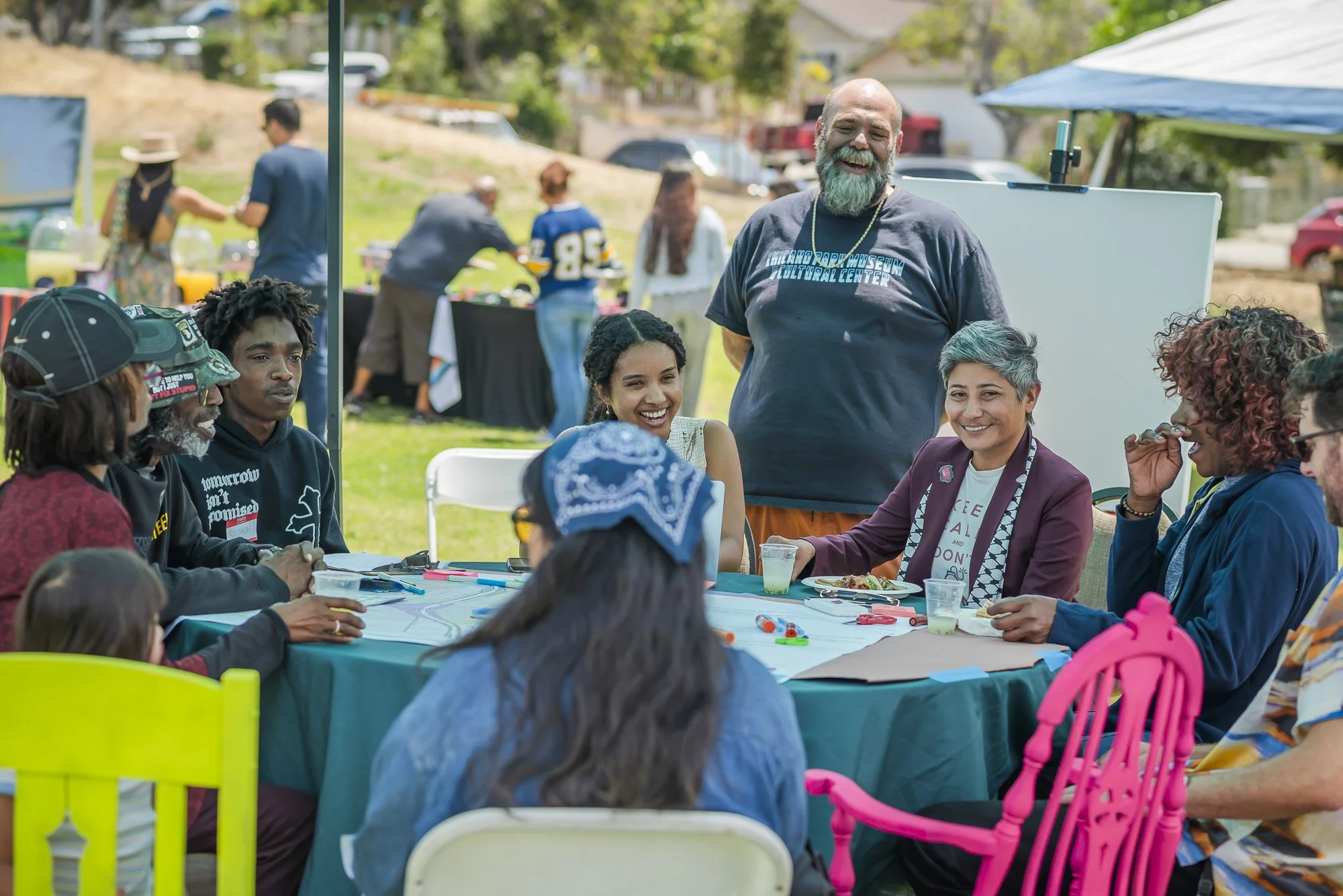 A diverse group of people gathered around a table outdoors at a community event, engaging in conversation and eating. In the background, other attendees are setting up booths, and there are trees and parked cars.
