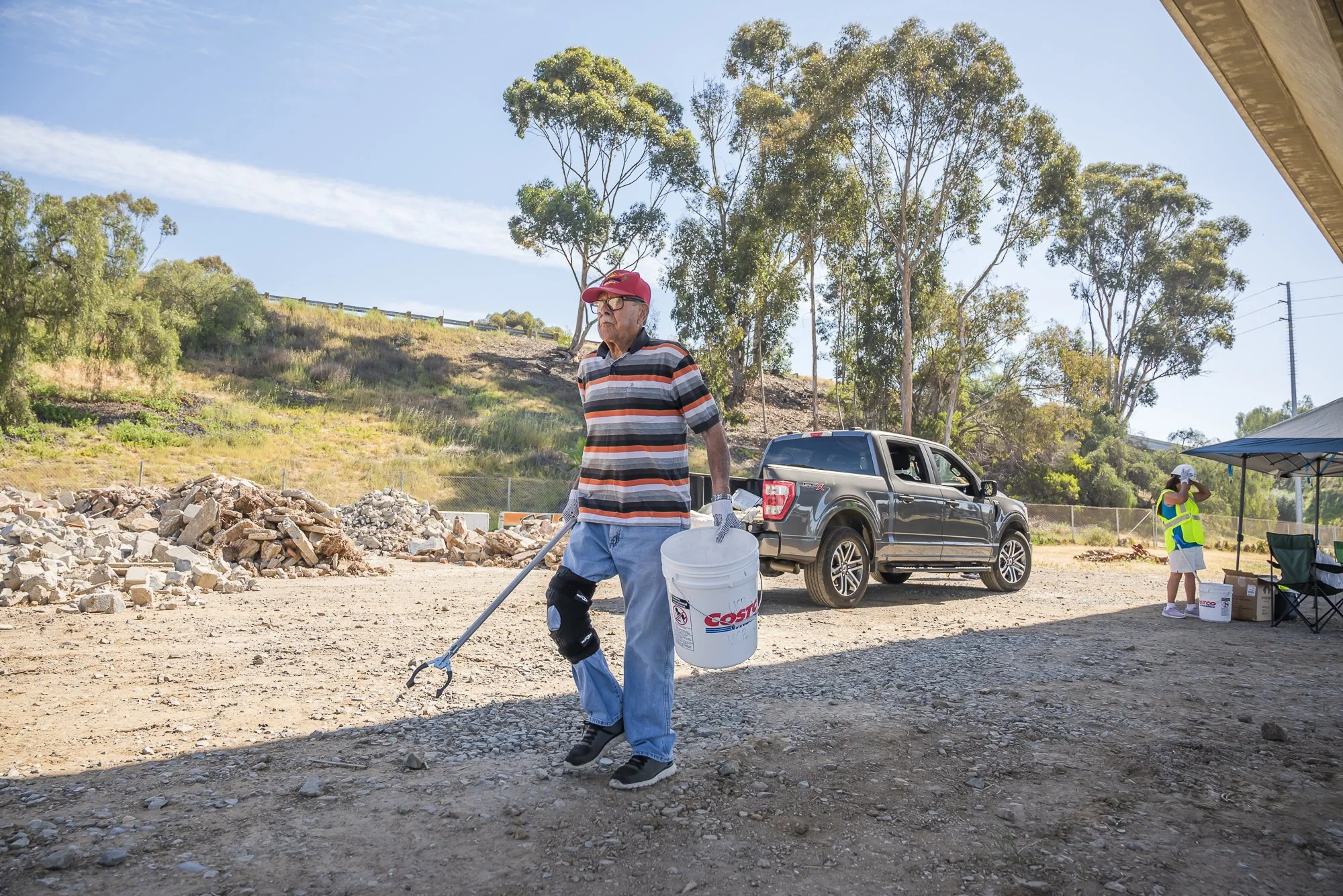 Man in striped shirt and cap carrying a white bucket, standing near a pile of rocks at an outdoor construction or landscaping site, with a silver truck and a field of trees in the background.