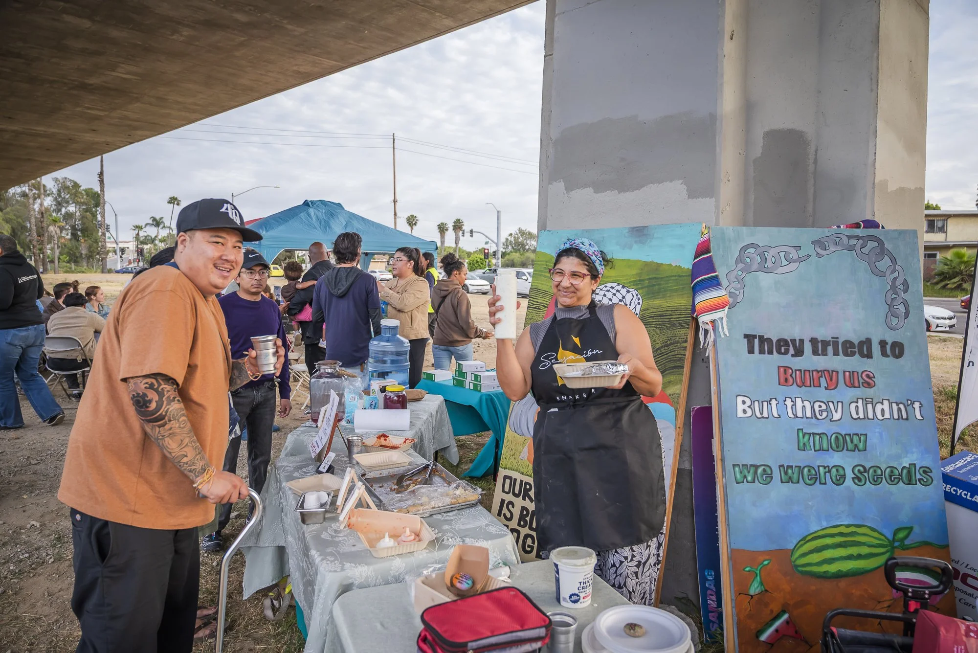 A group of people at an outdoor event under a bridge with a table of food and drinks. A woman holds a drink and wears glasses and a headscarf, smiling. A man with tattoos on his arm and a baseball cap stands nearby. Other people are socializing in th