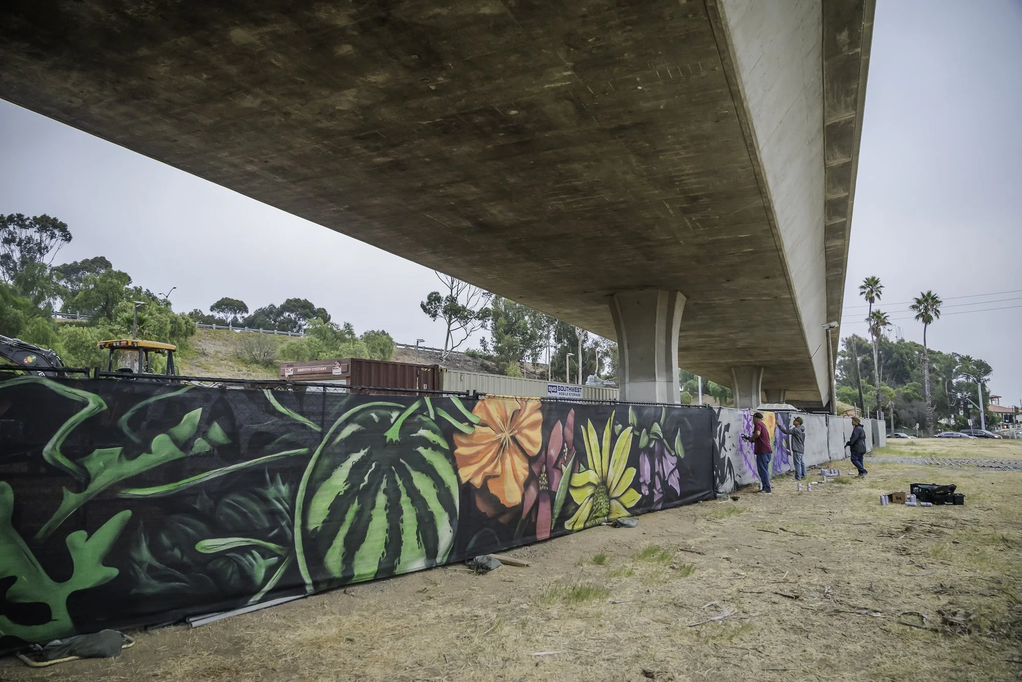 Underpass with concrete structure and support columns, a chain-link fence painted with colorful flower and leaf graffiti, a few people standing near the fence, trees and a clear sky in the background.