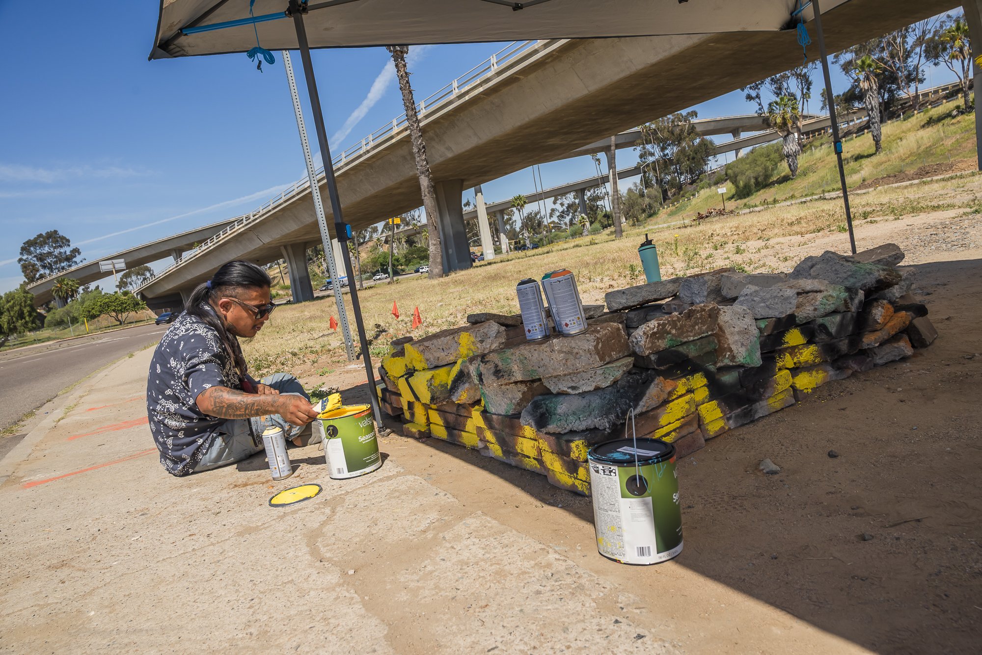 A person with long dark hair, tattoos, sunglasses, and a black patterned shirt is painting graffiti on a pile of large rocks with spray paint cans. They are kneeling on the sidewalk outdoors under a canopy with a bridge, trees, and a grassy area in t